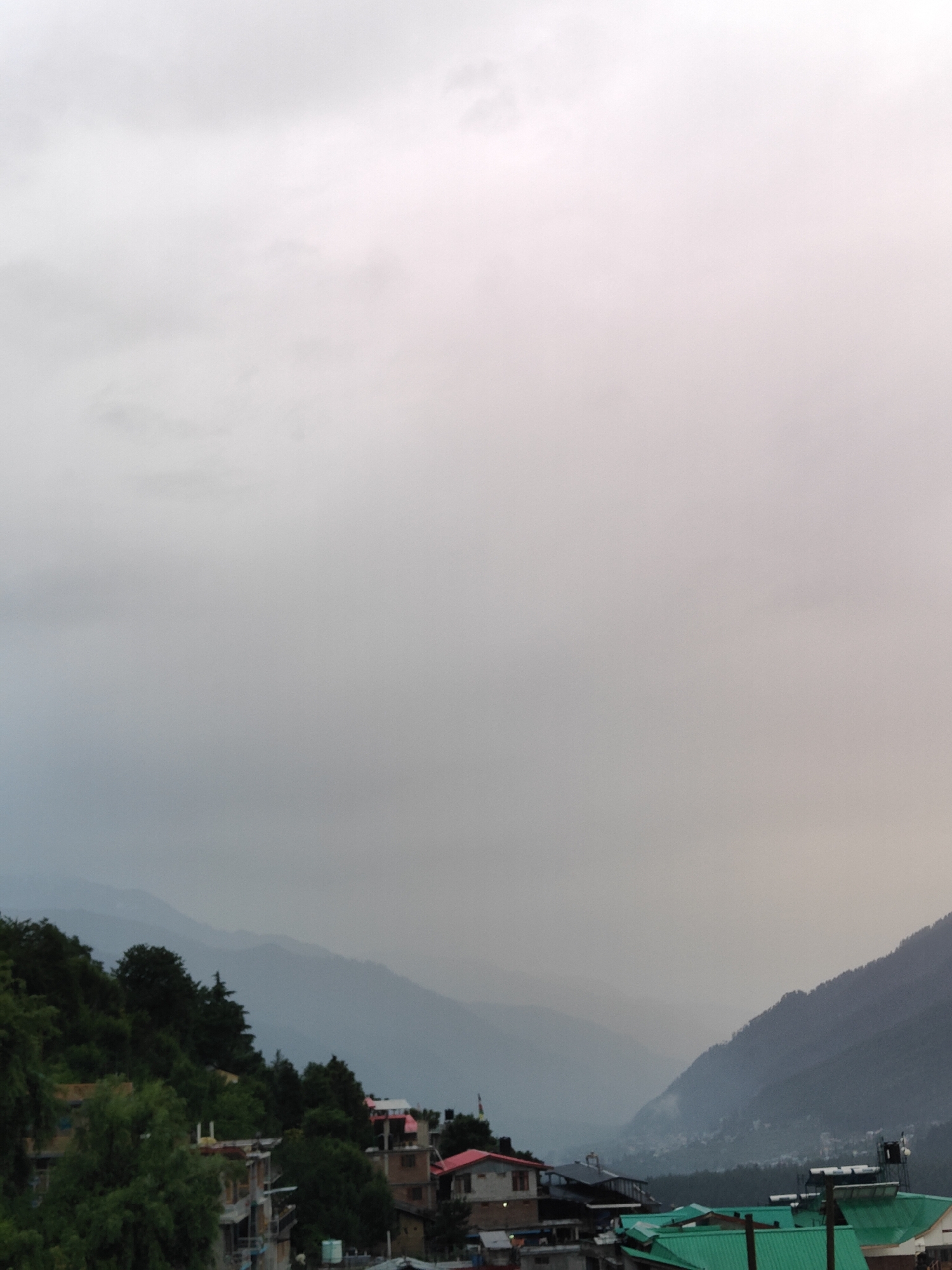 The image shows a mountain village in Vashisht, India, with buildings nestled on a hillside. The sky is overcast with a hazy, diffused light, suggesting it could be early morning, late afternoon, or a cloudy day. Lush green trees and foliage cover parts of the landscape, contrasting with the architecture. The buildings appear to be a mix of traditional and modern construction, with some featuring red roofs and others with green tin roofing. Solar water heaters are visible on some rooftops, indicating the use of renewable energy. The overall impression is one of a tranquil, mountainous settlement.