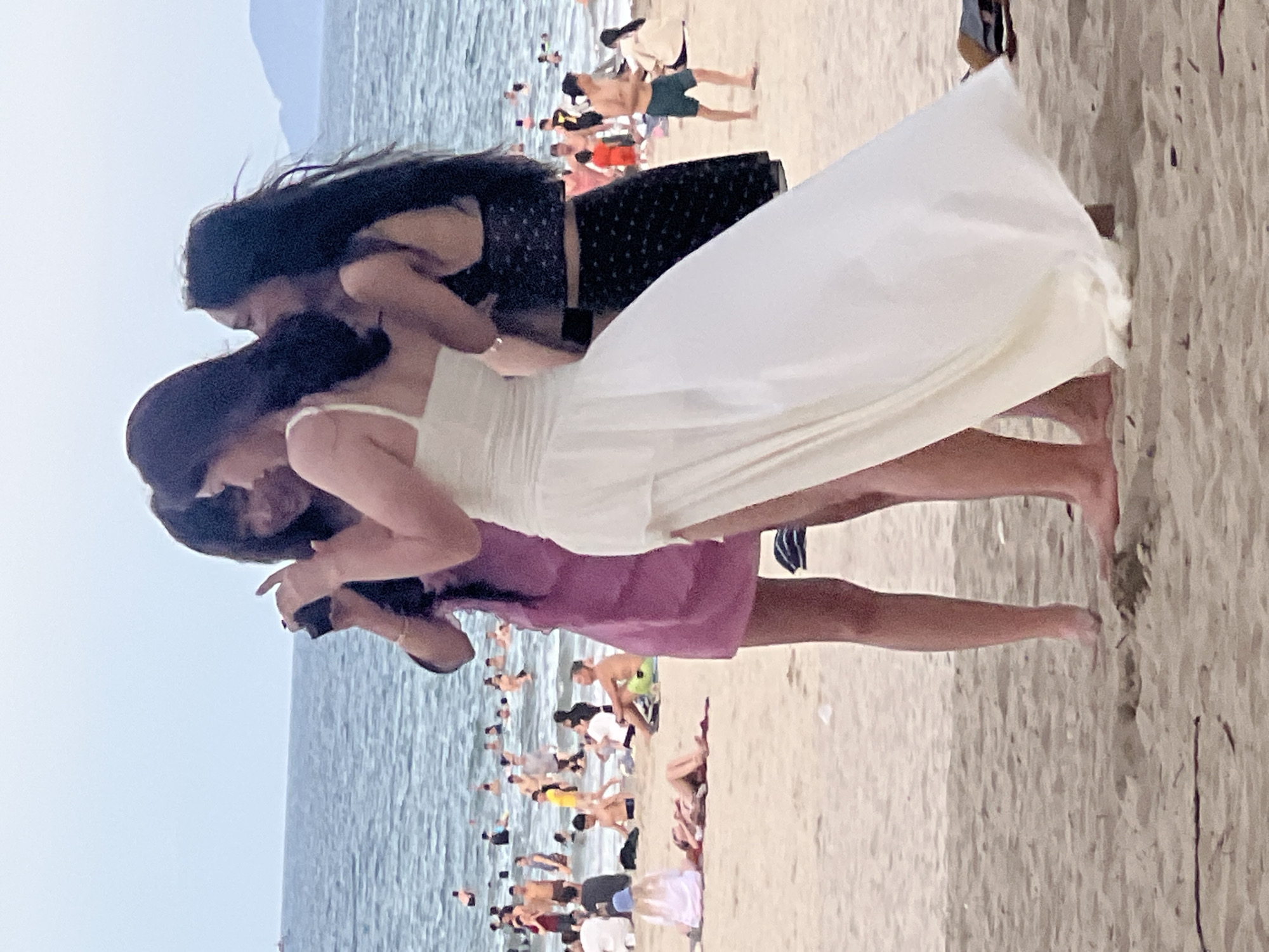 This is a vertical, somewhat blurry, medium-angle, eye-level shot of a crowded beach in Đà Nẵng, Vietnam. The foreground is dominated by three young women, two of whom are embracing while a third stands behind them. The woman in the center is wearing a white dress, the woman to her left has dark hair and is wearing a dark patterned top and skirt, and the woman on the right is wearing a purple dress. One of the women is holding a phone and appears to be taking a selfie.
The background shows a clear, bright sky and a body of water that appears to be the ocean, with many people scattered along the shoreline and in the shallow water. The beach is sandy and appears to be quite populated with individuals and small groups. The overall impression is of a sunny, active day at the beach. No text is visible in the image.