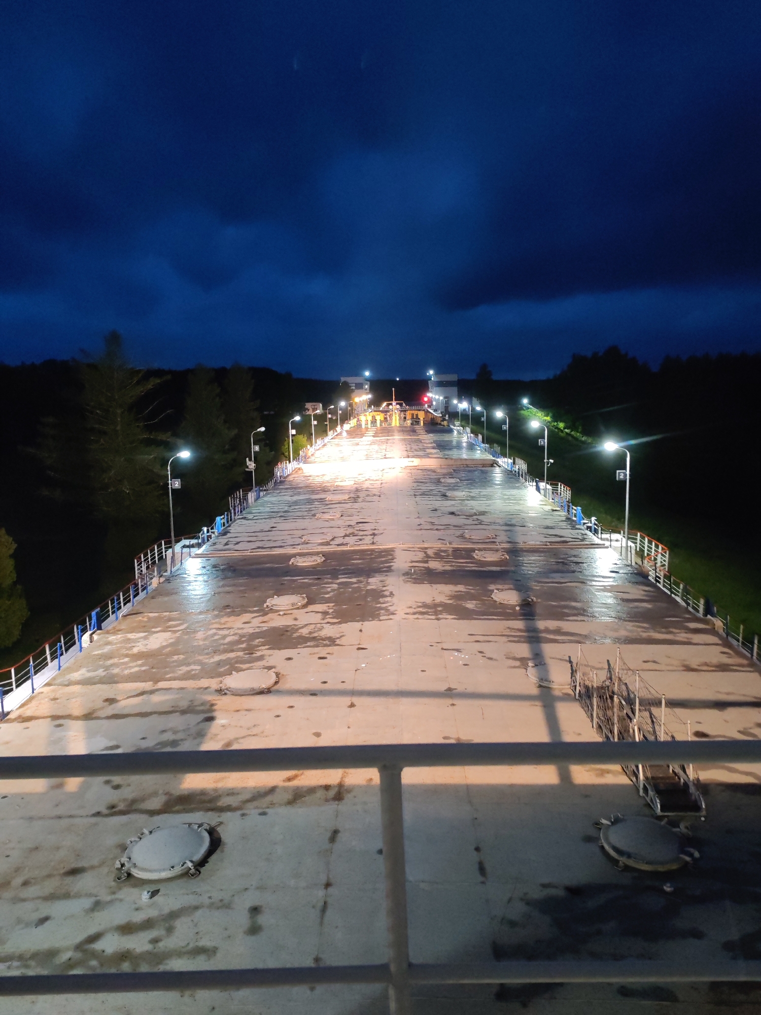 The image shows a long, wide concrete pier or walkway at night, illuminated by streetlights that line both sides. The walkway is wet, reflecting the lights, and has several circular manhole covers visible on its surface. The pier extends into the distance towards a brighter area with structures and traffic lights, possibly an entrance or gate. On either side of the pier, there are metal railings, and beyond the railings, dark trees and grassy slopes can be seen, indicating a natural or semi-natural environment. The sky is dark blue with dramatic clouds. The location is specified as Devyatiny, Russia. There are no people or discernible actions taking place in the scene.