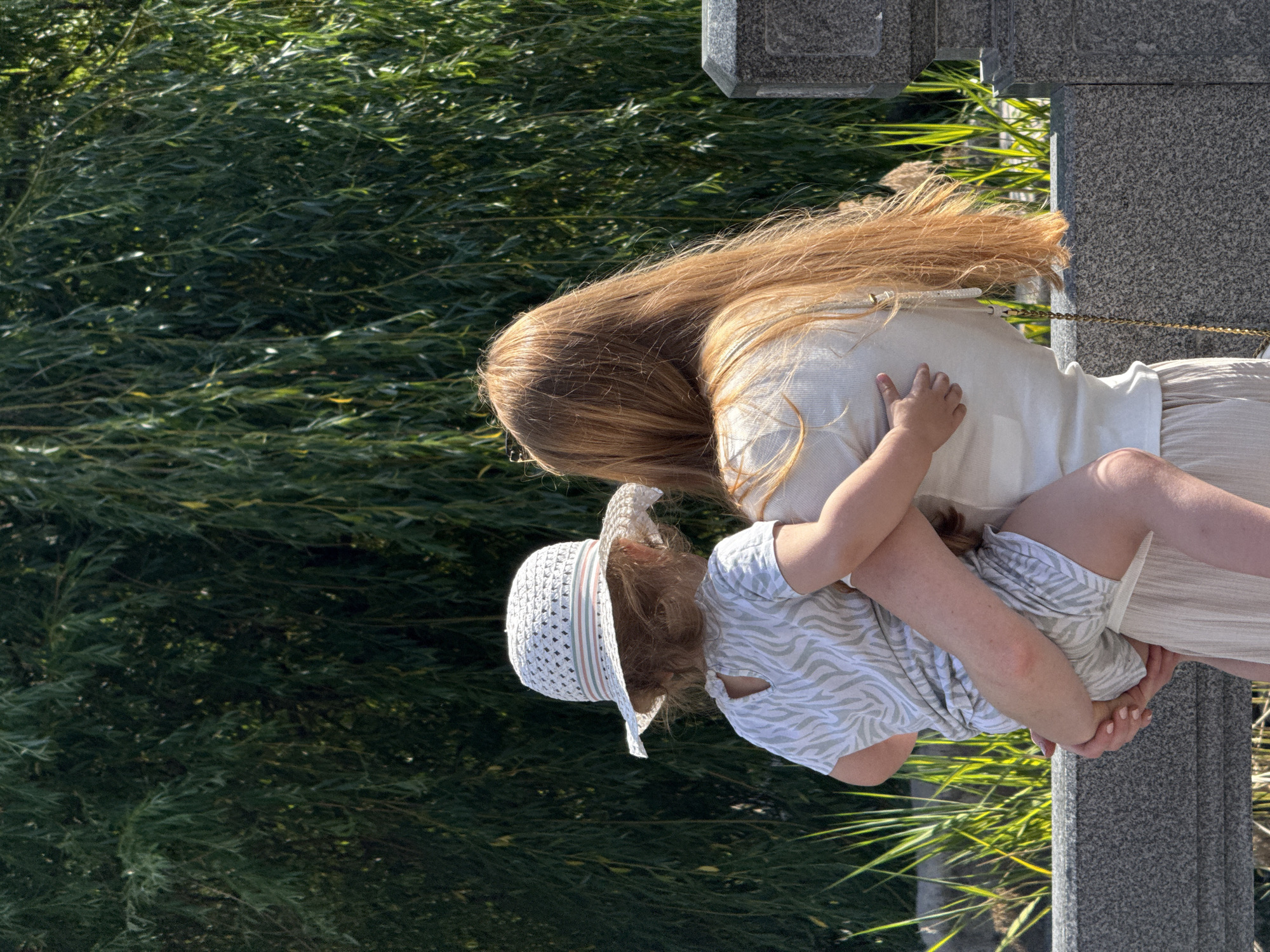 A woman with long, red hair is holding a toddler, who is wearing a straw hat and a zebra-print outfit, in her arms. They are standing near a stone railing, with a dense background of weeping willow trees and tall grass. The scene appears to be outdoors on a sunny day. The location is unknown, but it is unlikely to be Moscow, Russia, as there are no identifying landmarks or features indicative of that city.