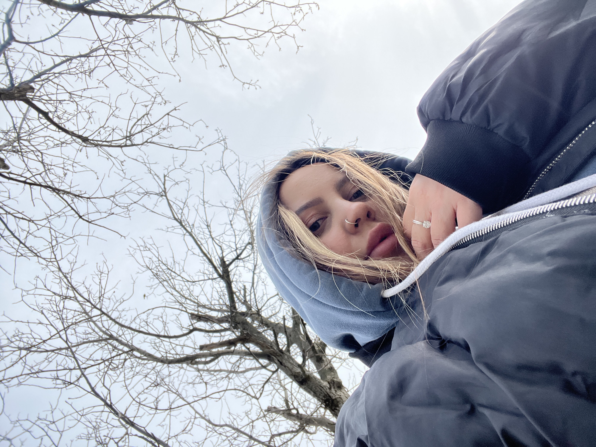 A low-angle selfie of a young person with blonde hair and a nose ring, wearing a blue hoodie under a black puffer jacket. The person is holding their hand up with a ring on their finger. The background consists of bare tree branches against a cloudy sky. There is no definitive evidence within the image to confirm the location is Novopokrovskaya, Russia.