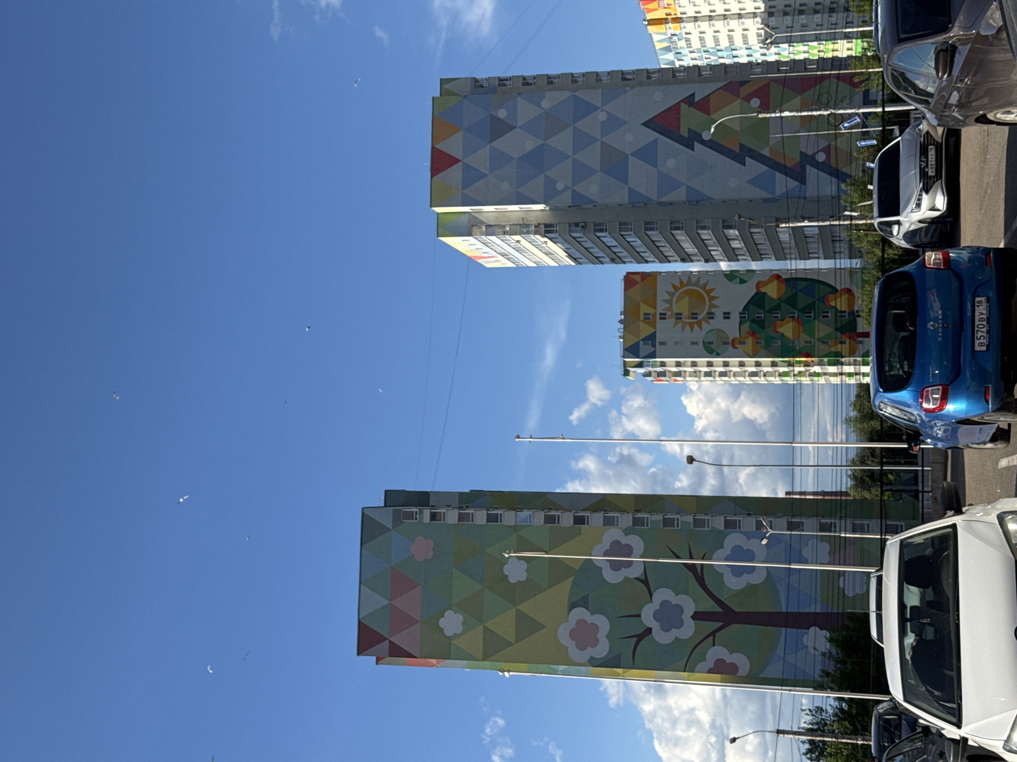 The image shows a row of apartment buildings in Izhevsk, Russia, decorated with large murals. The buildings are painted with geometric patterns, trees, and a sun. Cars are parked along the street next to the buildings. The sky is blue with scattered clouds. There are several birds flying in the sky.