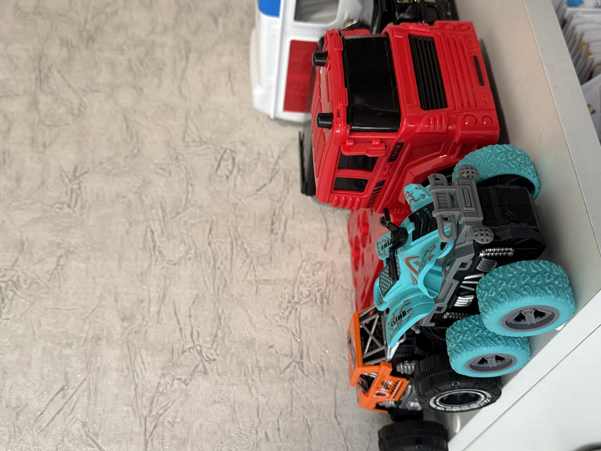 In the foreground, a bright blue and gray toy all-terrain vehicle with teal wheels is positioned on a white shelf. To its left and slightly behind, an orange toy off-road truck is visible. Behind the ATV, a red toy truck is prominently displayed. Further back, a portion of a white toy vehicle with a red and blue stripe is partially visible. The background consists of a textured, light-colored wall. The scene appears to be an indoor setting, likely a shelf displaying children's toys. The location is not discernible from the image itself, and therefore cannot be specified as being in Яромінскі сельскі Савет, Belarus.