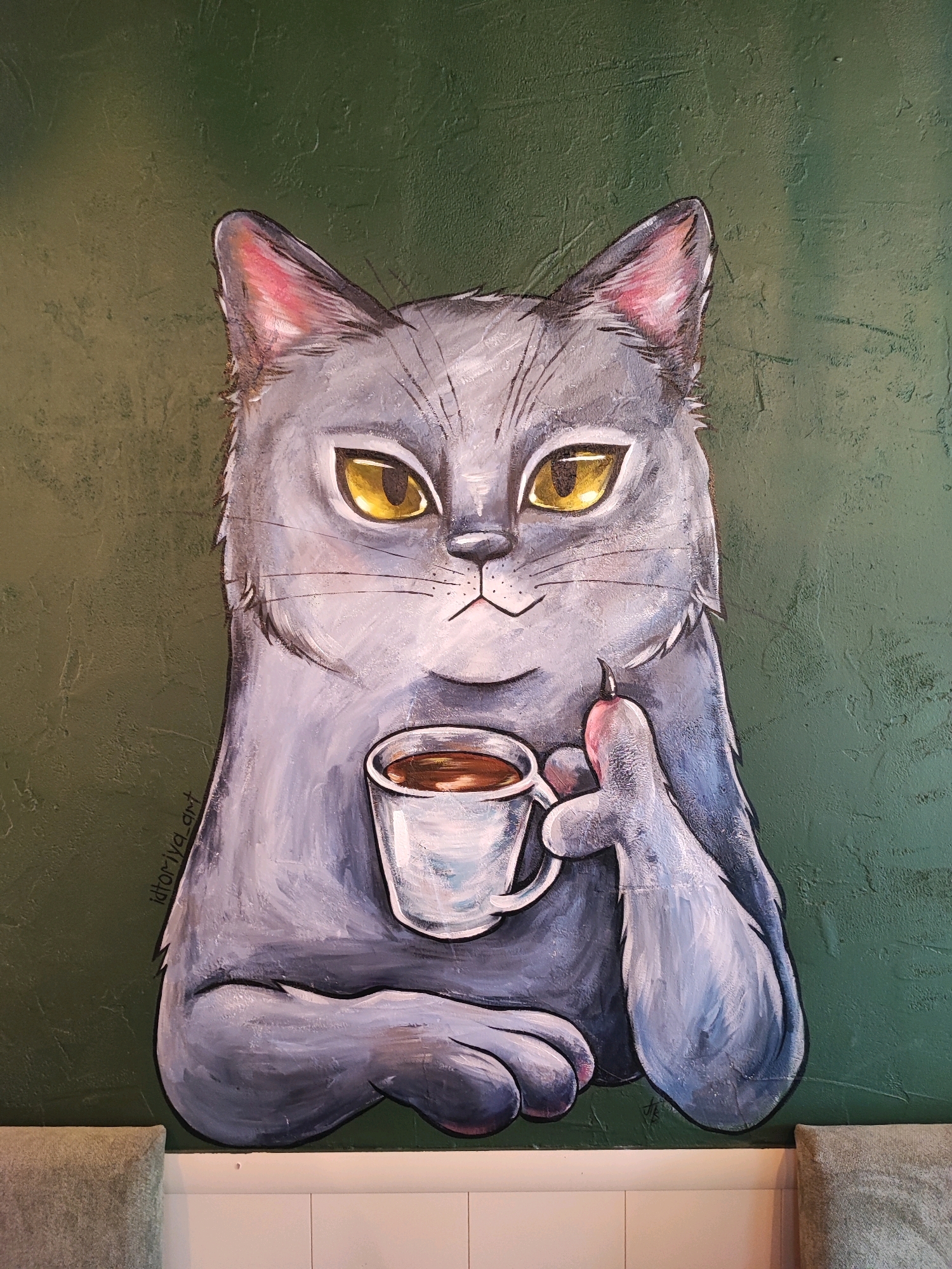 This is a painting of a grey cat with yellow eyes holding a cup of coffee and a lit cigarette. The cat is depicted with its body facing forward, but its head is turned slightly to the left, giving it a curious expression. The background of the painting is a textured green wall. A signature that appears to read "Idtoriya_art" is visible on the left side of the painting, and another smaller signature appears on the bottom right. This artwork is located on the wall of the cafe "Coffee House" on Lermontov Street in Pyatigorsk, Russia. The bottom of the image shows parts of upholstered furniture and a white tiled wall.