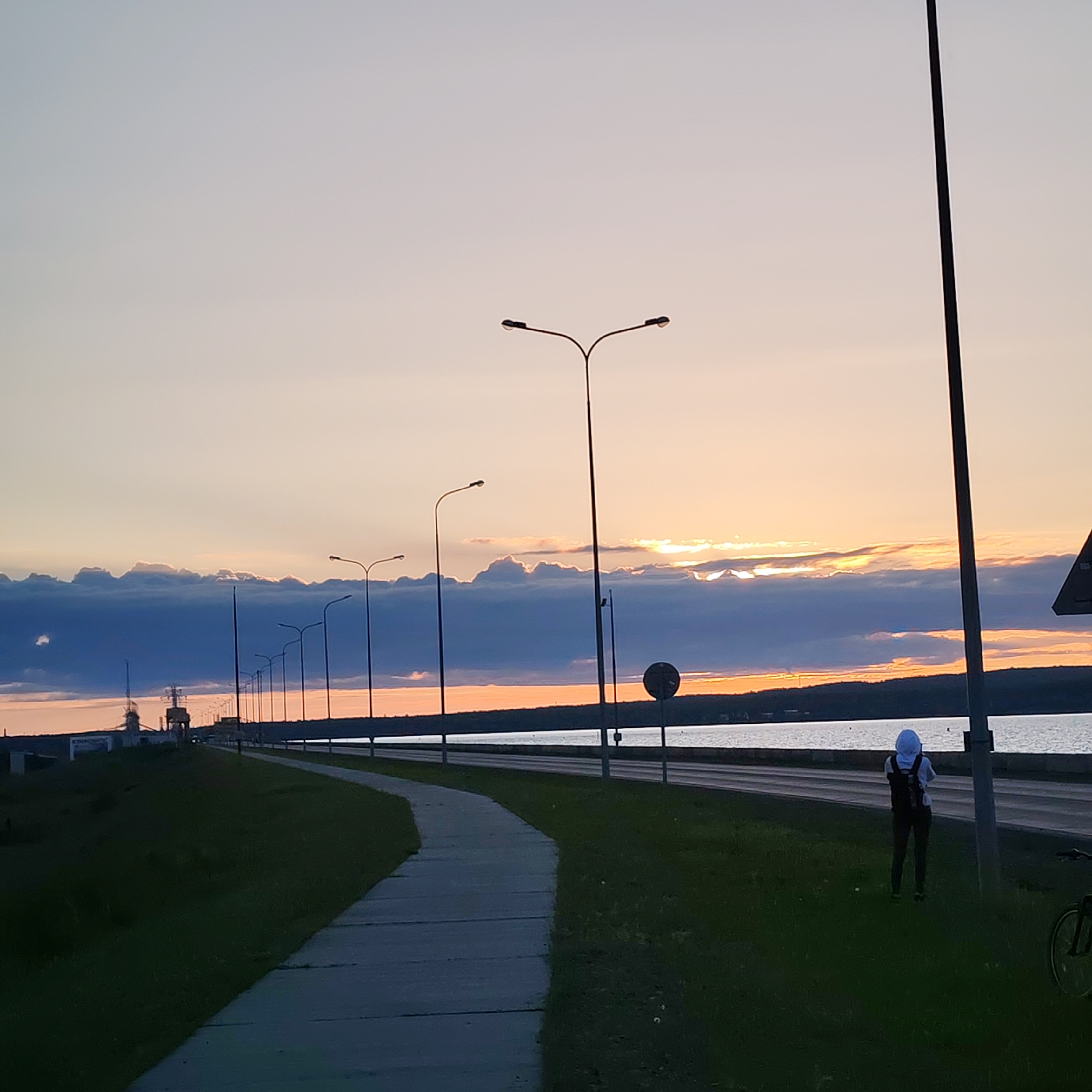 This image depicts a tranquil sunset scene alongside a large body of water in Chaikovsky, Russia. The time of day is clearly sunset, with the sky showcasing a beautiful gradient of colors, transitioning from light grey-blue at the top to soft oranges and yellows near the horizon where the sun is low. A prominent band of dark, layered clouds stretches across the mid-sky, with some sunlight breaking through, creating bright patches.
In the foreground, a paved pathway curves gently to the left, bordered by a strip of green grass. Parallel to the path on the right is a road, also following the contour of the water's edge. Numerous tall, dark streetlights with dual lamps line the road, silhouetted against the bright sky.
One person is visible on the grassy area between the path and the road, standing with their back to the camera. They are wearing a white hooded garment or head covering, dark pants, and a dark backpack, appearing to be observing the sunset over the water. A dark, circular road sign is partially visible near the person. In the bottom right corner, a portion of a bicycle wheel and frame can be seen.
The calm water reflects the soft light of the sky. Across the water, a dark, forested landmass forms the distant shoreline. To the far left, what appear to be industrial structures or port cranes are faintly visible against the horizon. The scene evokes a peaceful, contemplative atmosphere. There is no visible text in the image.