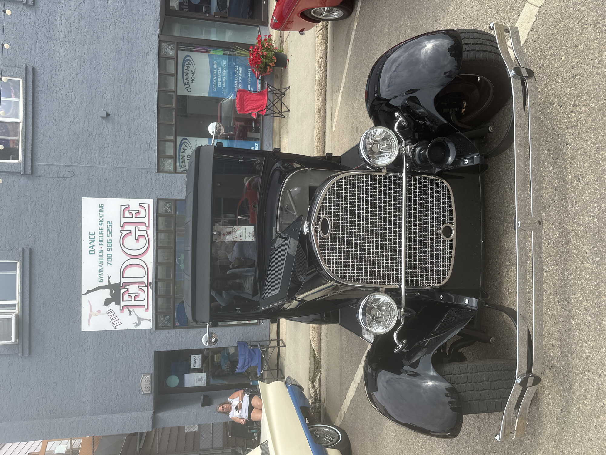 A vintage black Ford Model A coupe is parked on a paved street in front of a building with businesses named "The EDGE" dance studio and "CLEAN MY HOME" cleaning services. The building is grey and has the address 4816B visible above a closed door. A woman sits in a chair outside the studio, looking at her phone. Red chairs and potted flowers are visible outside the cleaning service business. The car has a chrome grille, round headlights, and a visible horn. The background shows more businesses and a glimpse of other vehicles. The scene appears to be in the City of Leduc, Alberta, Canada, based on the phone number 780-986-5252 displayed on "The EDGE" sign, which is a local area code for Leduc.