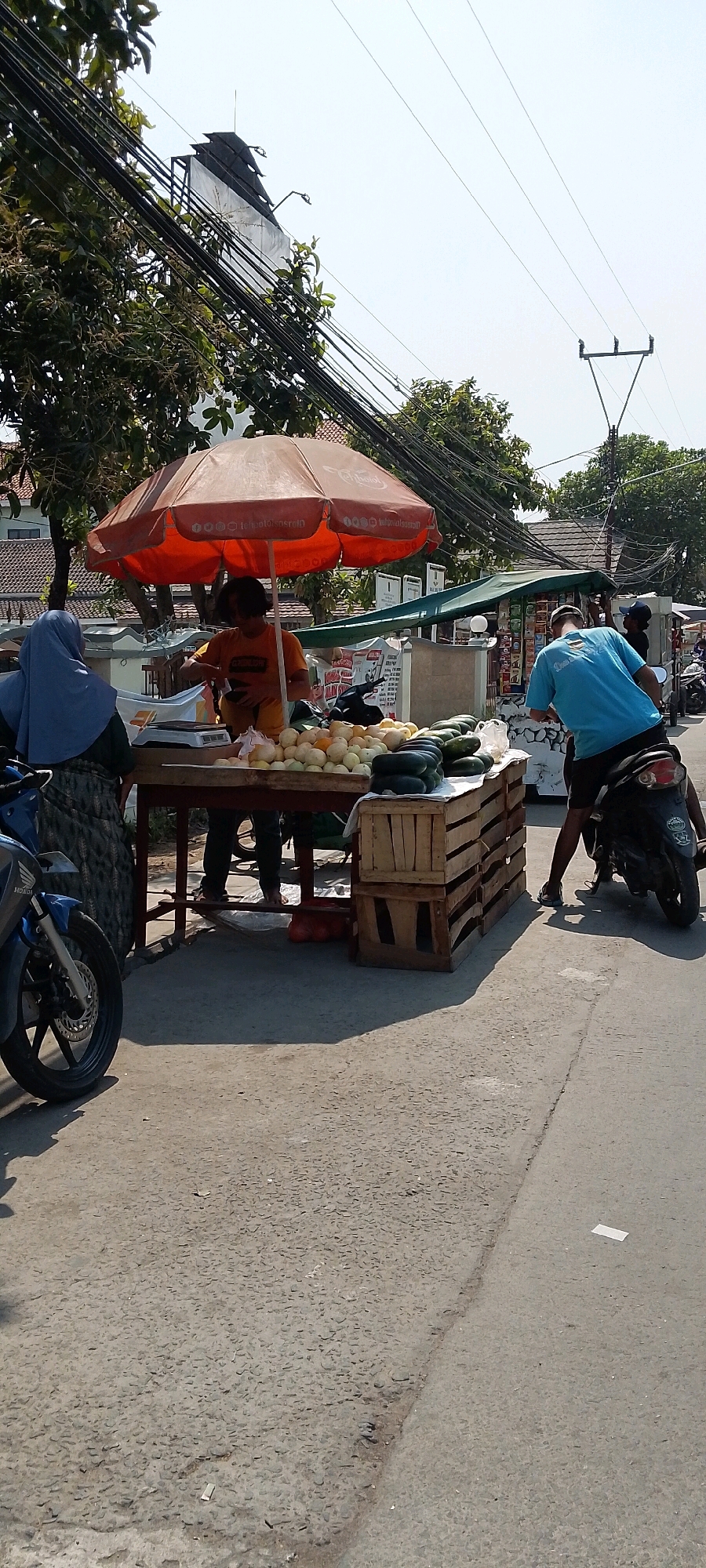 In Babelan Kota, Indonesia, on a sunny day, a street vendor is selling produce under an orange umbrella. A customer, wearing a blue hijab, stands to the left, facing the vendor. The vendor, in an orange shirt, is attending to a customer. A variety of fruits and vegetables are displayed on a wooden table and in wooden crates, including melons, cucumbers, and other items. A motorcycle is parked to the left of the stall. In the background, there are buildings, trees, and a complex network of overhead electrical wires. Another person on a motorcycle is in the process of turning, with their back to the camera, and is wearing a blue t-shirt. The scene takes place on a paved street.