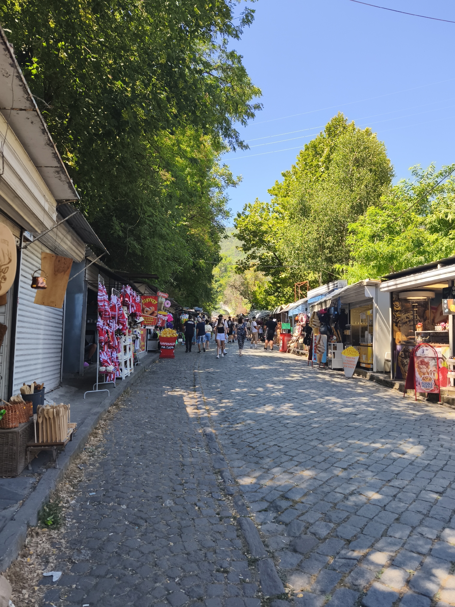 The image depicts a cobblestone street lined with small market stalls on both sides. The stalls on the left appear to sell souvenirs, with racks of red and white embroidered items and wooden crafts visible. On the right, some stalls seem to be selling food or drinks, with ice cream cones and signs suggesting confectionery items. Several people are walking along the street, browsing the stalls. Lush green trees overhang the street from both sides, casting dappled shadows on the cobblestones. The sky is clear and blue. The scene suggests a lively pedestrian area, likely a tourist or market district, within the city of Bachkovo, Bulgaria.