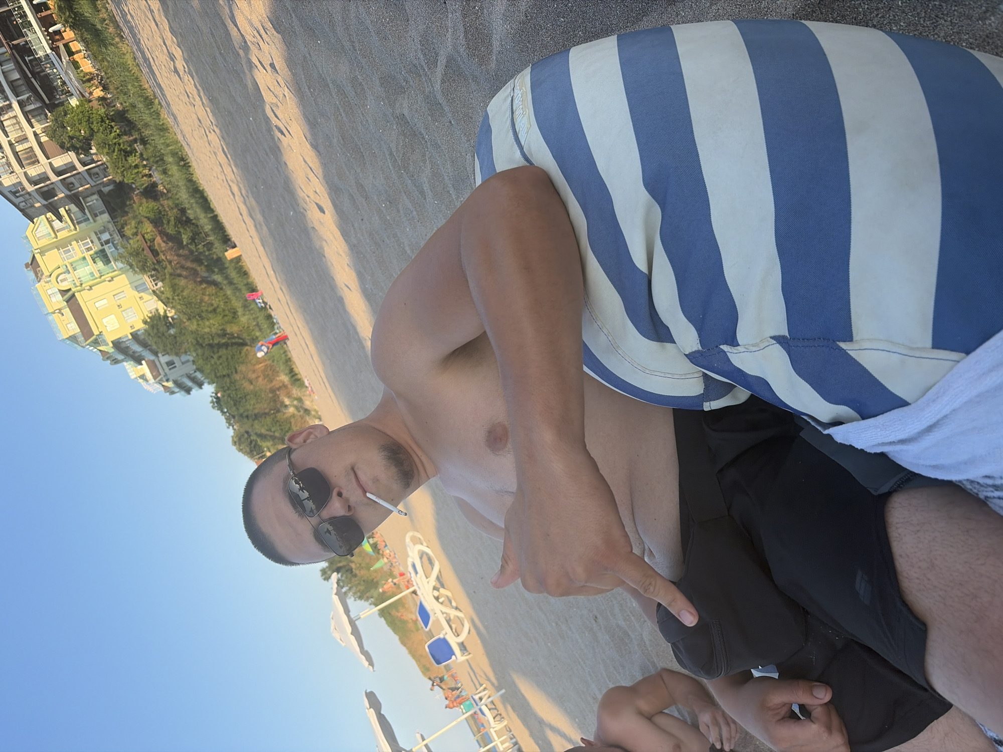 The image shows a man sitting on a sandy beach in Lozenets, Bulgaria, with buildings and trees on a hillside in the background. The man is shirtless, wearing sunglasses and black shorts, and is smoking a cigarette. He is giving a thumbs-up and pointing. He is sitting on a blue and white striped cushion. There are beach chairs and umbrellas visible in the middle ground, and other people are scattered on the beach in the background. The sky is clear and blue.