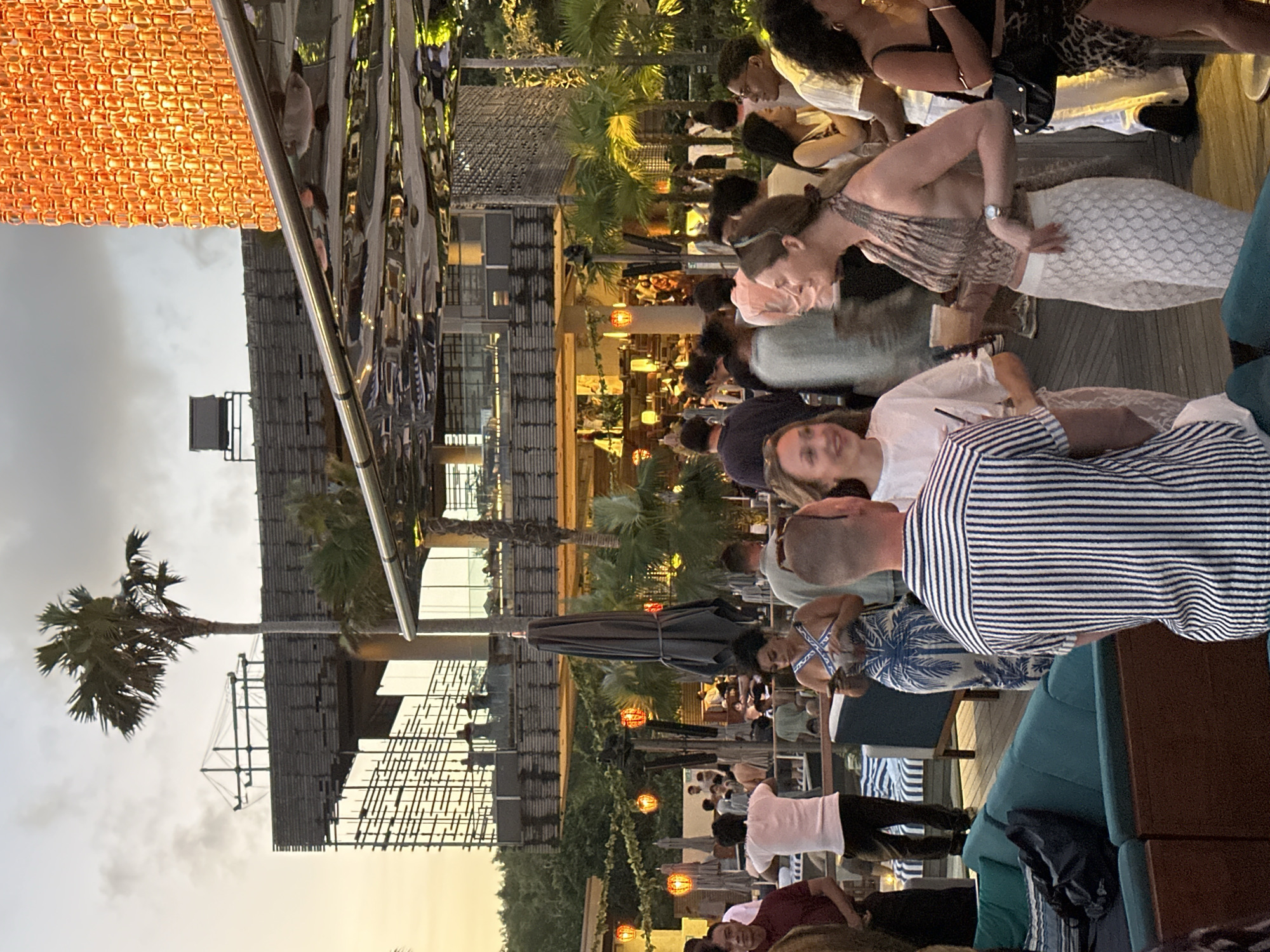 The image depicts a busy outdoor setting, possibly a rooftop bar or restaurant, with numerous people socializing. The location is likely in Kuta Selatan, Indonesia, given the presence of tropical vegetation such as palm trees and the architectural style of the building. People are dressed in casual clothing, with some standing and mingling while others are seated at tables. The building appears to be multi-storied with a modern design, incorporating latticework and reflective surfaces. Decorative elements, such as hanging lights, add to the ambiance. Some individuals are close to the camera, allowing details like clothing patterns (e.g., stripes, floral prints) to be observed. The lighting suggests it's either dusk or early evening.