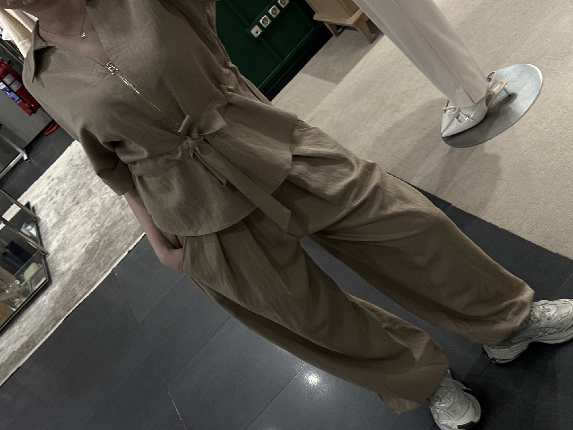 The image shows a person wearing a tan-colored linen two-piece outfit, consisting of a top with a zipper and a tied waist, and loose-fitting pants. The person is also wearing white sneakers. The setting appears to be inside a store with a mix of carpeted and dark tiled flooring. There is a display stand with a mannequin wearing white clothing and white pointed-toe heels visible. Behind the person, there is a green cabinet and a display case with glass shelves. The location is unknown, and there is no indication in the image that it is in Sofia, Bulgaria.