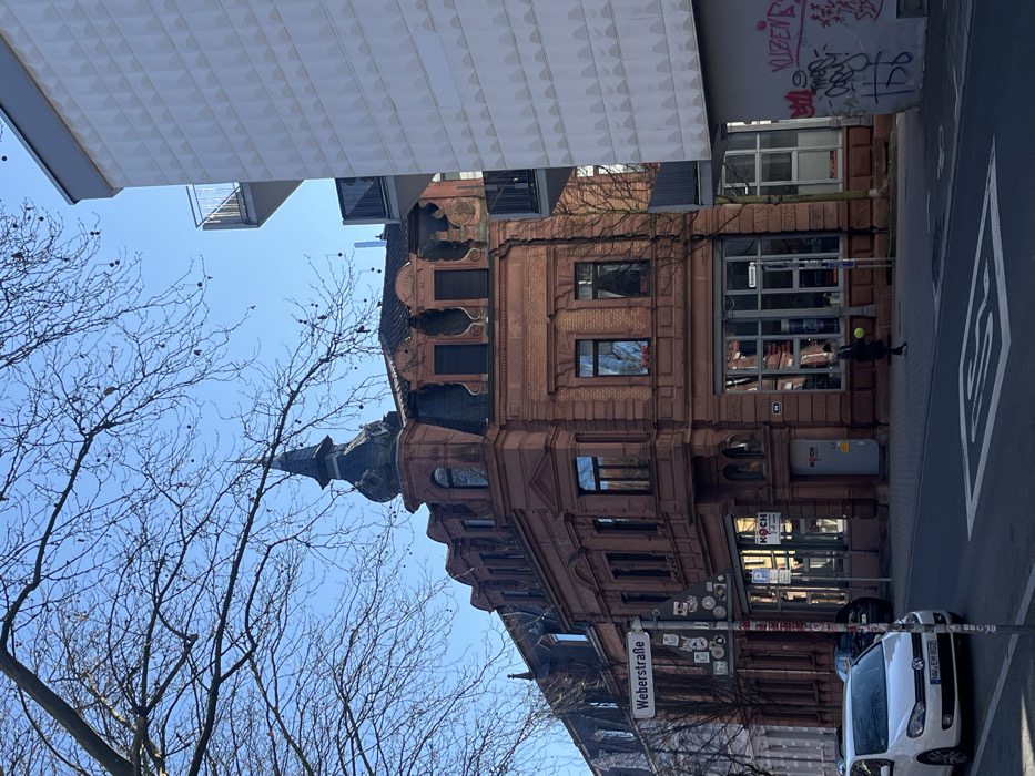 The image depicts an urban street scene at Weberstraße in Kaiserslautern, Germany, under a clear blue sky. Bare tree branches are visible in the upper portion of the frame.
The central feature is a multi-story building on a corner, constructed with a reddish-brown brick facade and a dark gray mansard roof. It features an ornate corner tower with a spire. The ground floor includes large display windows and an entrance with a "KOCH" sign. Adjacent to this building on the right is a modern, multi-story structure with a light-colored, textured facade and dark gray balconies with metal railings. The lower section of this modern building's wall exhibits graffiti. An additional older building is partially visible to the left of the historic corner building.
A white Volkswagen Golf (license plate KL-EM 602) is parked on the left side of the street. White road markings, including an arrow and a crosshatch pattern, are visible on the asphalt. A street sign pole in the left foreground displays "Weberstraße" and various stickers.
A single individual, dressed in dark clothing, is observed walking on the sidewalk in front of the historic building. This person is holding a bright yellow, spherical object in their left hand.