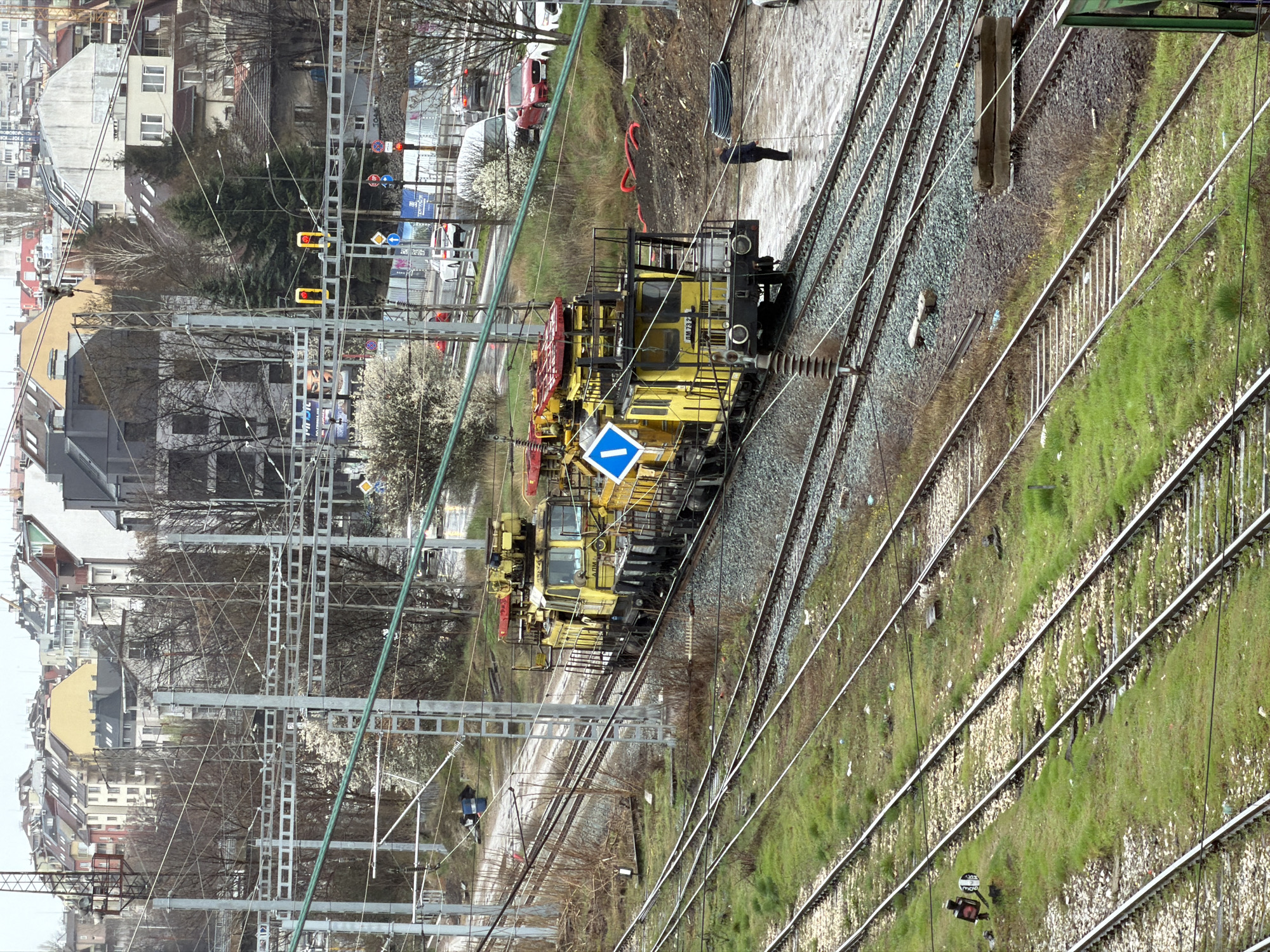 The image captures a scene in Sofia, Bulgaria, presenting a complex network of railway tracks in an urban environment. A specialized yellow railway maintenance train dominates the central view, positioned on the tracks. A blue directional sign is prominently displayed on the side of the train. The tracks themselves run parallel, interspersed with gravel and patches of green grass.
In the background, buildings characteristic of a European city can be observed, intermingled with bare trees and blossoming white trees, suggesting a transitional season. Railway signal lights are visible, adding to the complexity of the railway infrastructure. A pedestrian is walking near the tracks.