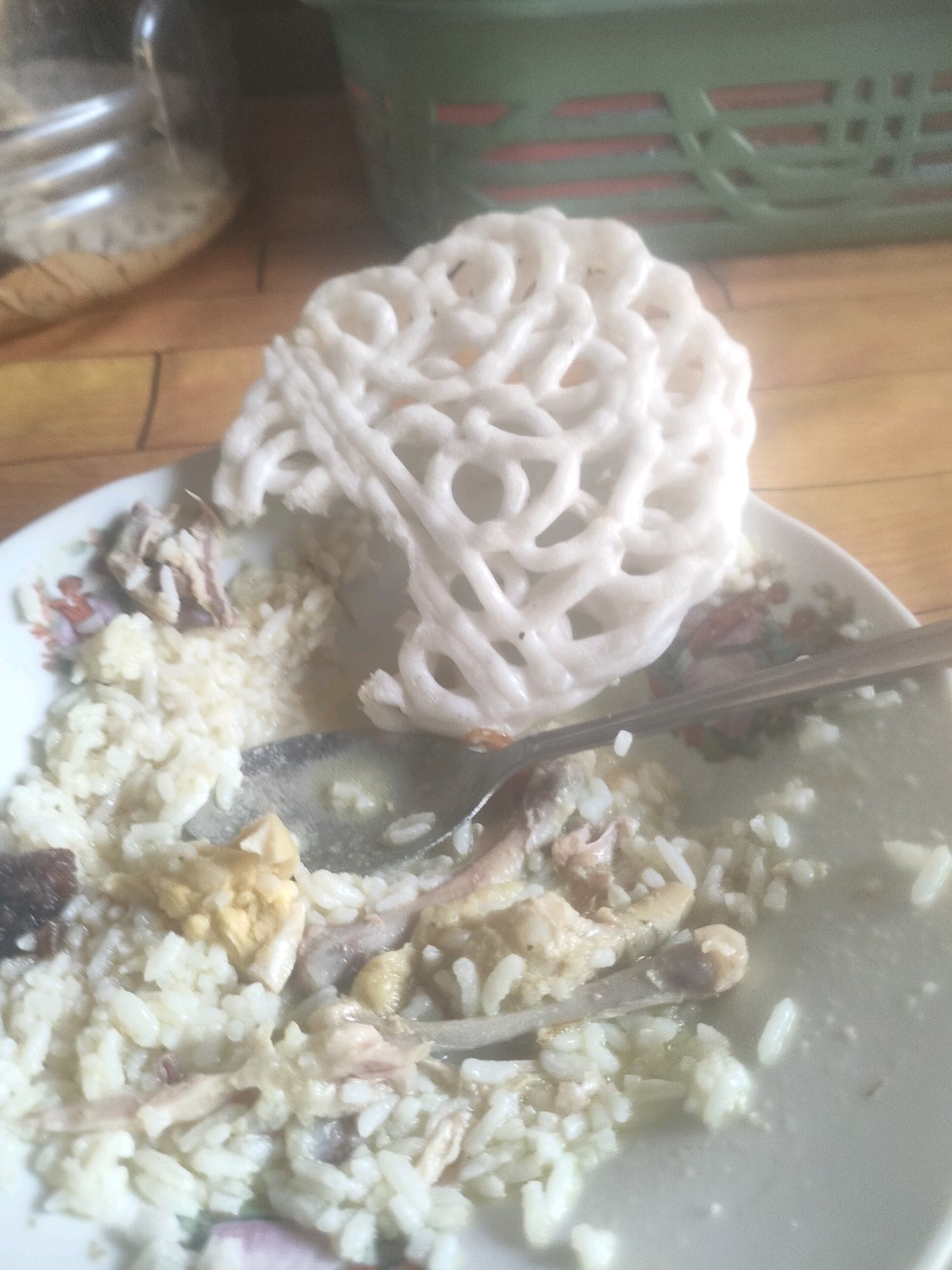 A close-up, slightly blurry, overhead shot shows a bowl of rice with chicken and a white cracker. The bowl is on a wooden table, with a blurred background that includes a glass jar and a green woven object. A spoon rests in the bowl.