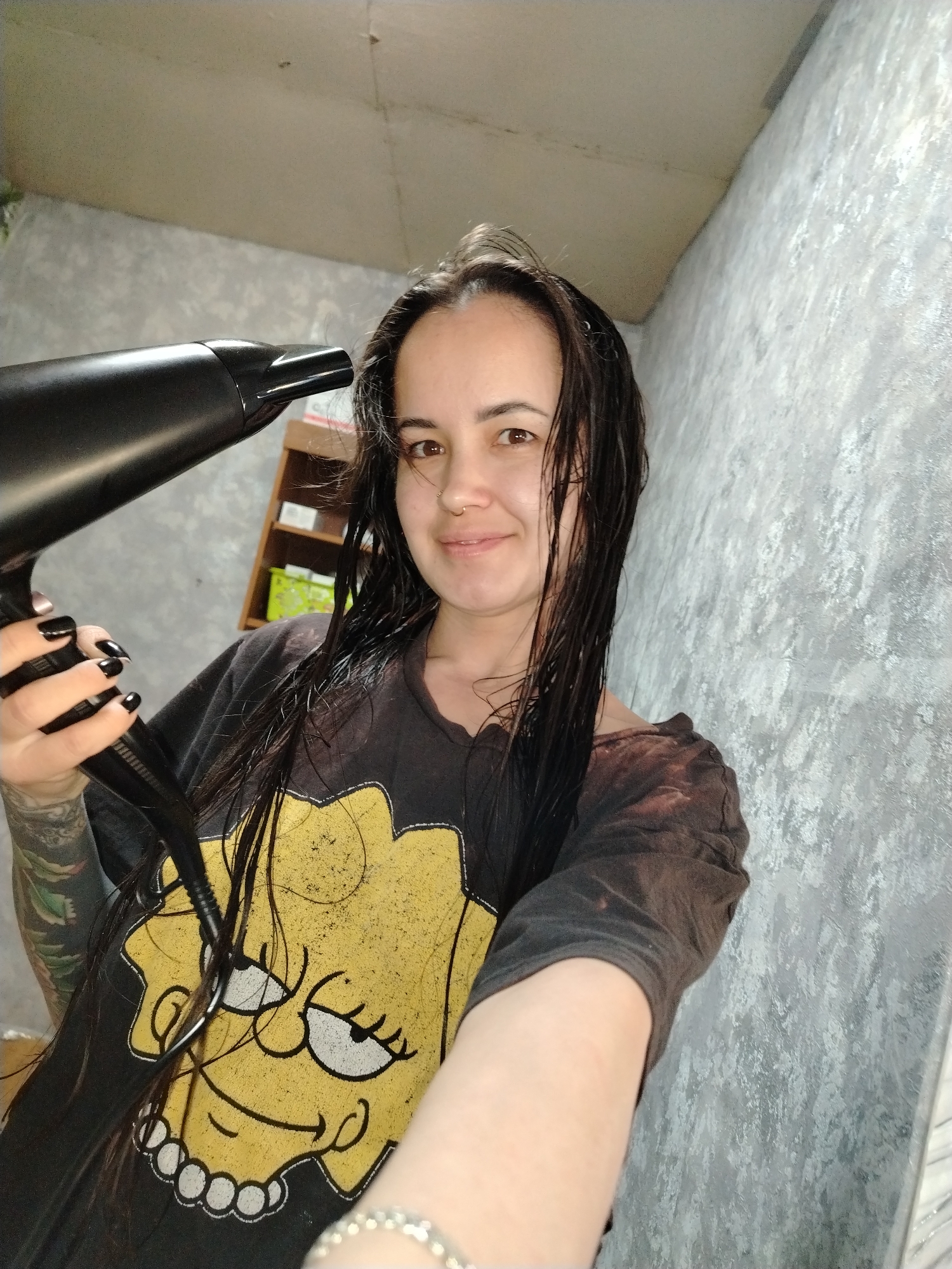 A young woman with dark hair is holding a black hair dryer in her right hand, pointing it towards the camera. Her hair is wet and plastered to her face. She is wearing a black t-shirt with a yellow cartoon character on it. She has a nose piercing. Her fingernails are painted black. A tattoo is visible on her left arm. In the background, there is a grey wall and a wooden shelf unit. The location is Гожан, Russia.