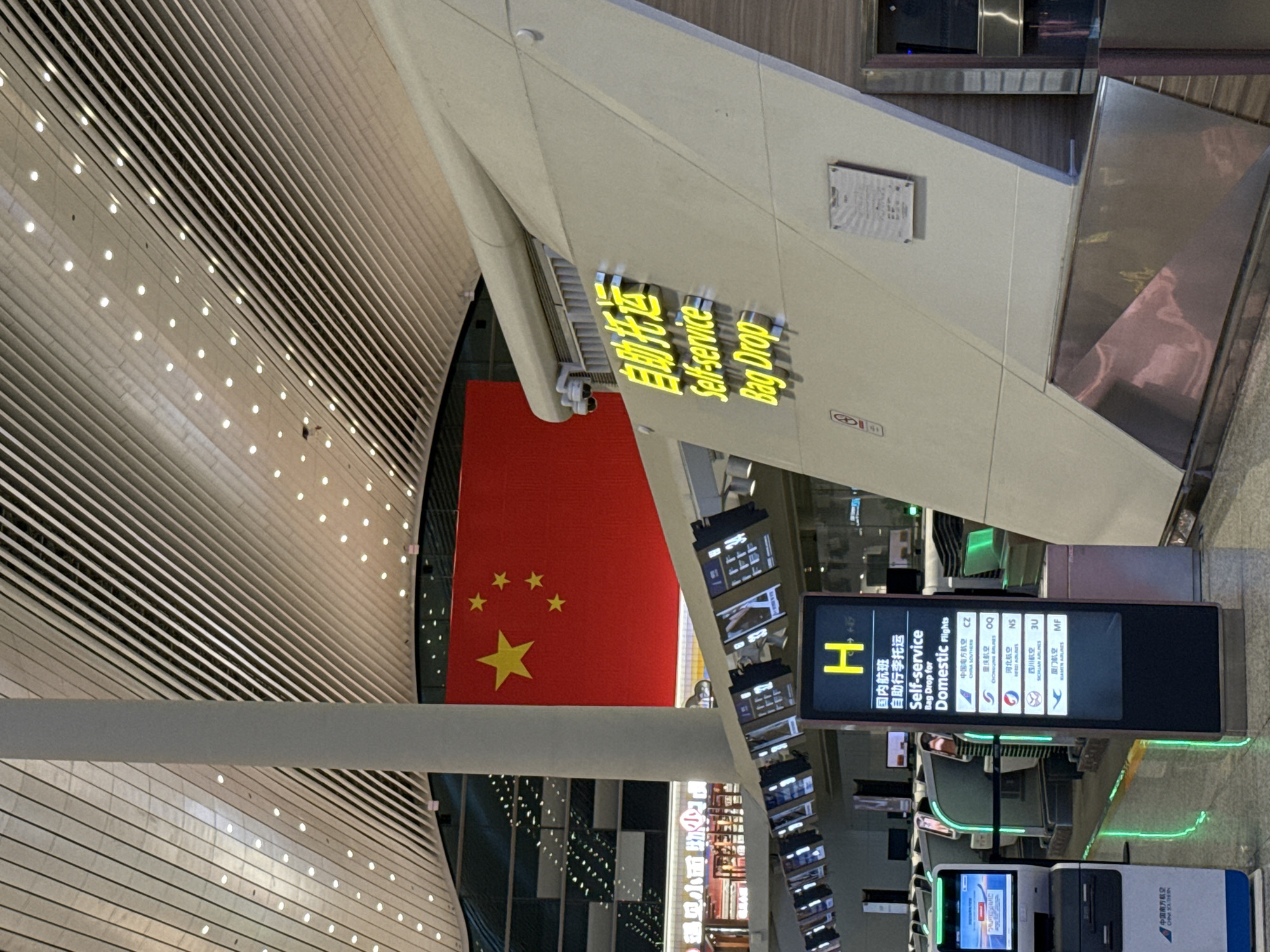The image shows an interior view of what appears to be the Guangzhou Baiyun International Airport in Guangzhou City, China. The foreground features a "Self-service Bag Drop" area, indicated by illuminated signage in both Chinese and English. A large screen displays information about domestic flights, listing airlines like China Southern, Chongqing Airlines, Hebei Airlines, Sichuan Airlines, and Xiamen Airlines. Below the screen, self-service kiosks are visible. Above, in the background, the Chinese flag is prominently displayed. The architectural design includes a ceiling with linear patterns of white bars and dots of light. The image also shows shops and the word "遇见小面" written in Chinese characters, which translates to "Meet Noodle."