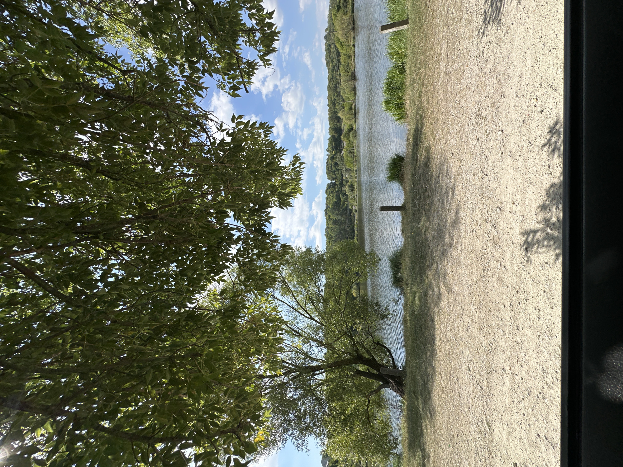 An outdoor scene shows a calm body of water, likely a lake or river, under a partly cloudy blue sky. The foreground is a dirt path, with tall grass and two wooden posts visible along the water's edge. A large tree with green foliage is situated on the left side of the image, its branches extending over the path and casting shadows. In the background, a line of trees and hills forms the horizon. The image appears to be taken from a shaded area, possibly under another tree or a structure, as evidenced by the dark horizontal band across the bottom and the dappled shadows. The overall impression is a natural, serene landscape. The location is identified as Makiv, Ukraine.