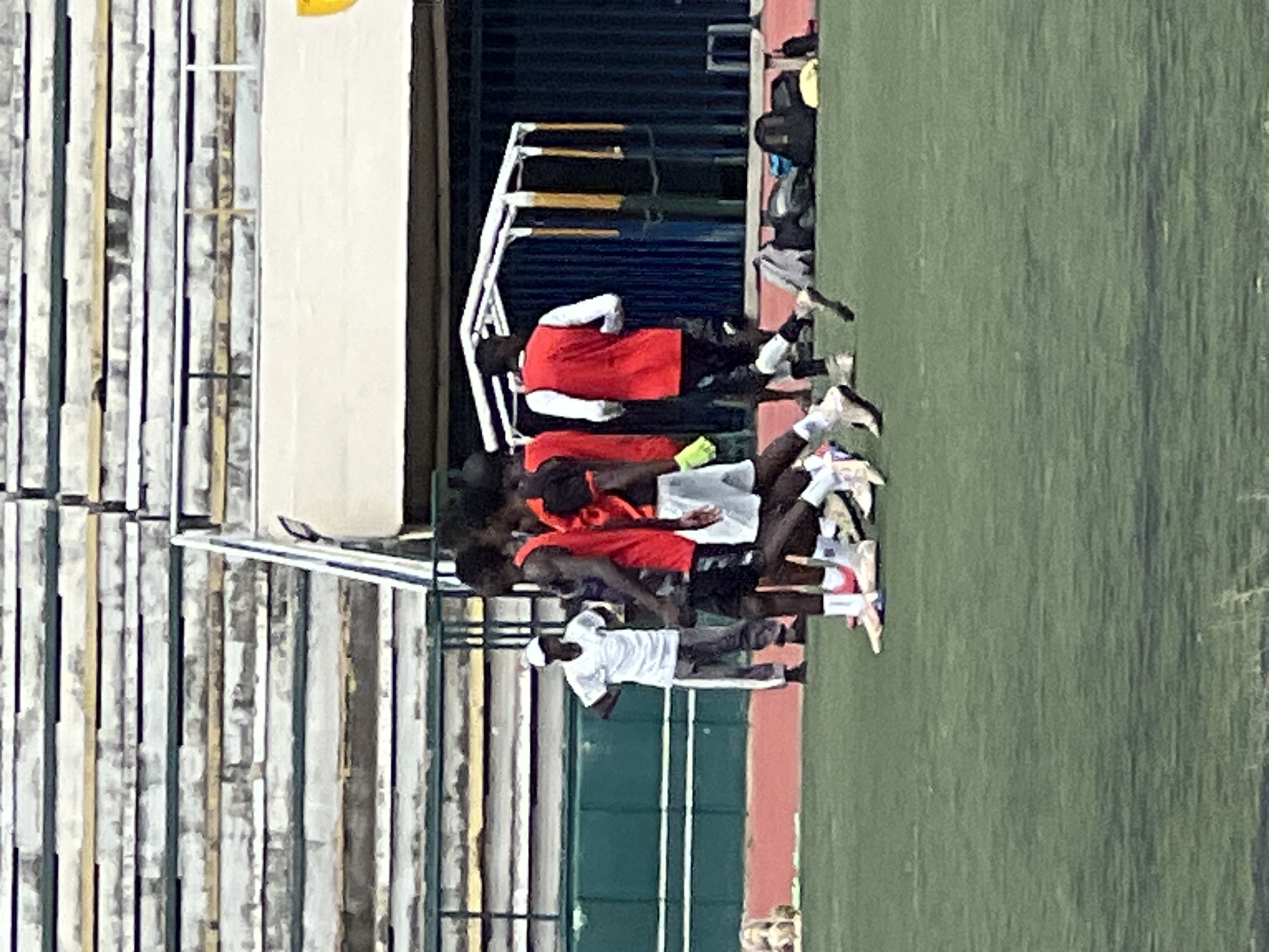 A group of young men in athletic attire are on a green field, possibly a soccer pitch, in front of tiered stadium seating. The stadium seating is weathered and appears to be made of concrete with yellow and green accents. In the background, there is a white tent-like structure with yellow poles and a dark blue curtain or wall behind it. A few bags and a person lying down are visible near a bench under the tent structure. One man in the foreground is wearing a white t-shirt and grey pants, standing with his hands on his hips. The other men are in athletic shorts and jerseys, some are walking or standing in a group. One man in the group wears a red jersey, another a black jersey with red trim. The lighting suggests it is daytime.