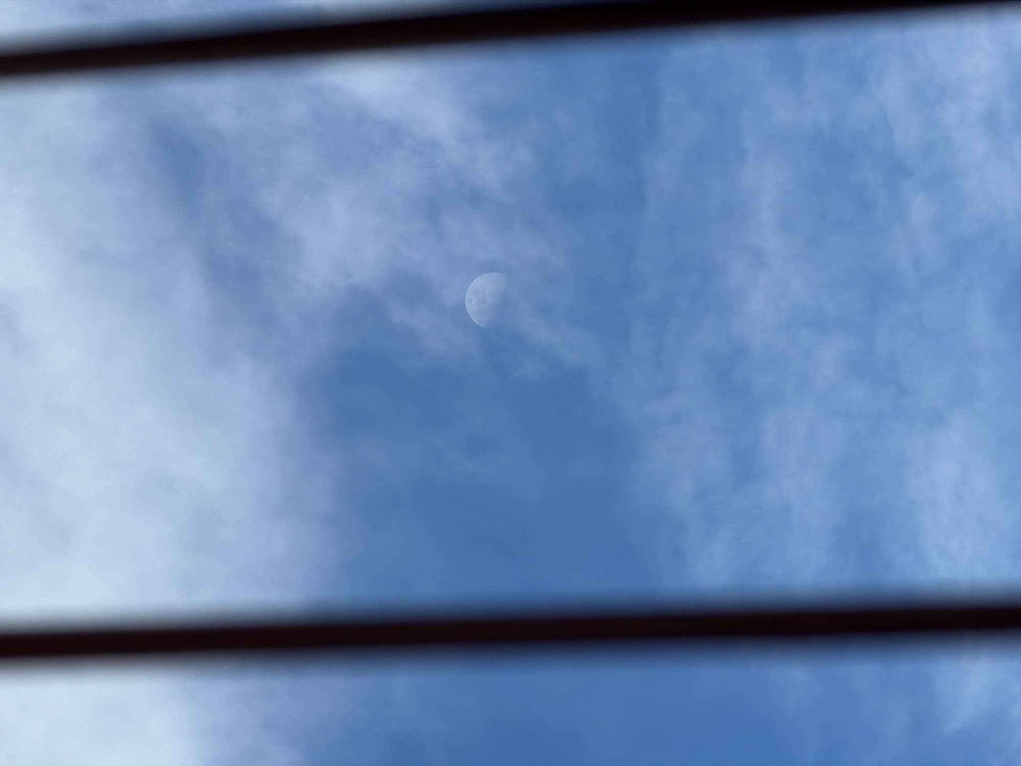 The scene depicts a daytime sky observed from Velingrad, Bulgaria. The view is partially obstructed by two dark, vertically oriented, out-of-focus foreground elements, appearing as bars or posts, flanking the left and right sides of the image. The sky is light blue, featuring scattered white and translucent grey cloud formations. A distinct waxing gibbous moon, approximately two-thirds illuminated, is visible in the upper-left quadrant, partially obscured by the ambient cloud cover. No persons, direct actions, or ground-level objects are present.