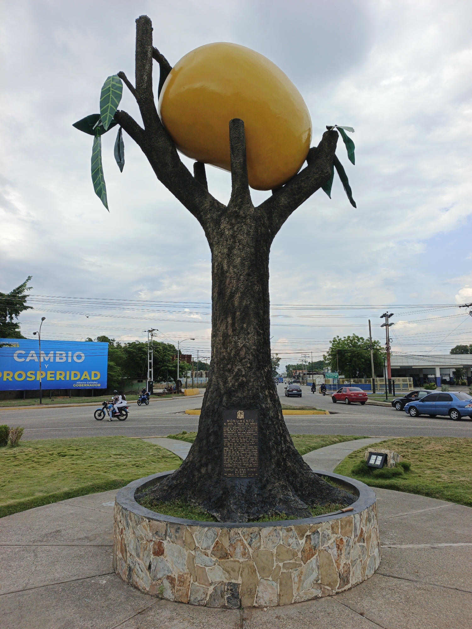The image shows the Monumento al Árbol y Fruto del Mango, a sculpture in San Carlos, Venezuela. The sculpture consists of a tree trunk with branches holding a large yellow mango. The trunk sits in a stone-clad circular base. A dark plaque is affixed to the trunk. The sculpture is on a traffic island with grass and pavement surrounding it.
In the background, a busy street is visible with vehicles, including cars and motorcycles. A large blue billboard advertising "Cambio y Prosperidad" is visible on the left side of the image. The sky is overcast with some blue patches. Other buildings and trees line the street in the background.