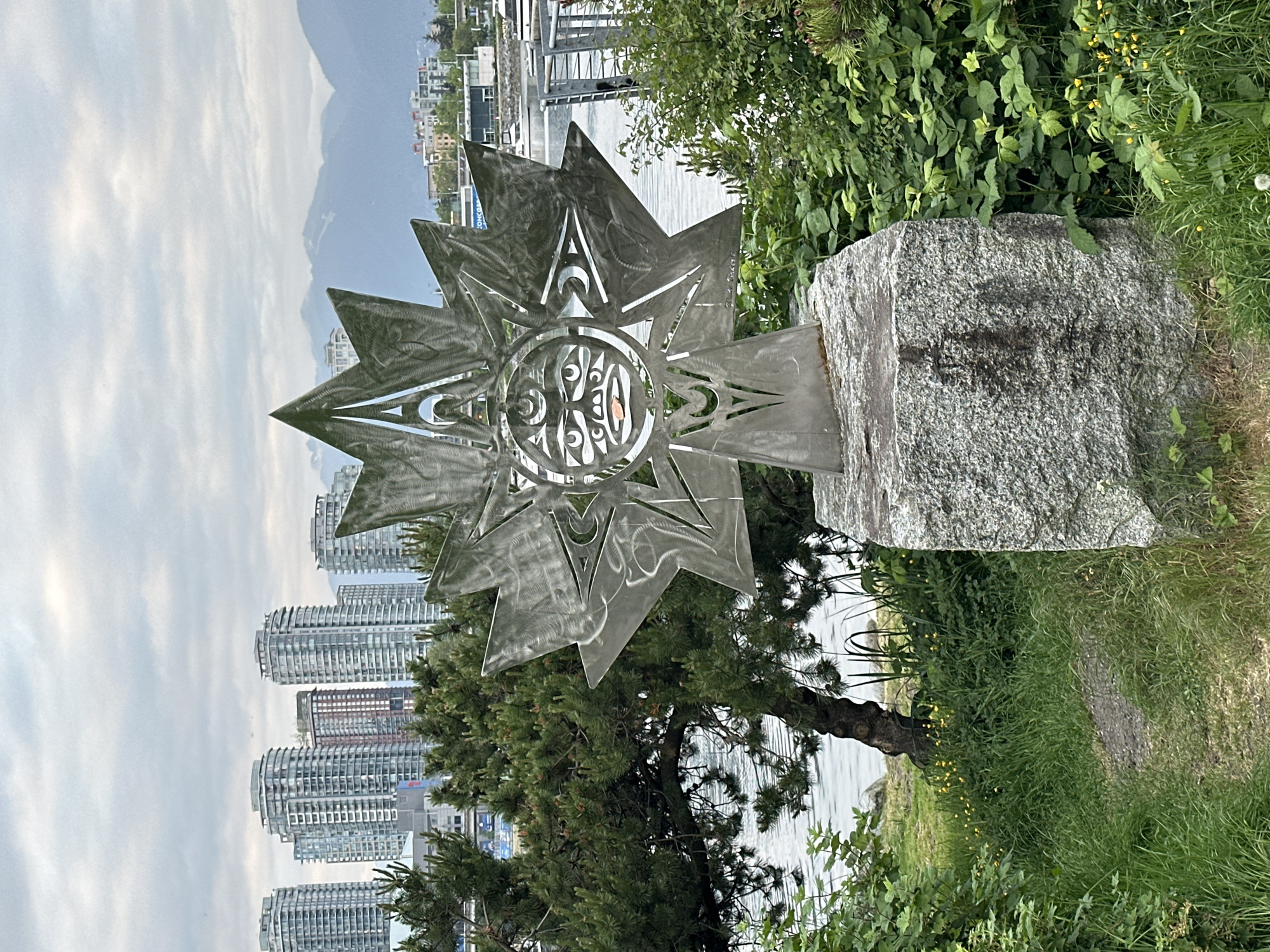 A landscape photograph depicts a metal sculpture set against a background of modern buildings, water, and a cloudy sky, indicating an urban waterfront location. The sculpture, which resembles a stylized sunburst or star with indigenous art motifs, is positioned on a large, grey granite-like boulder. To the right of the boulder, green bushes and grass form a natural border, while behind the sculpture, a pine tree bridges the space between the art piece and the urban skyline. A stretch of water runs horizontally across the midground, separating the sculpture and surrounding vegetation from the buildings. In the background, high-rise residential buildings with curved architectural designs dominate the skyline, suggesting a dense, urban environment. The sky is overcast with light grey clouds that diffuse the sunlight. The scene is situated in a park or public space within the city of Vancouver, Canada.