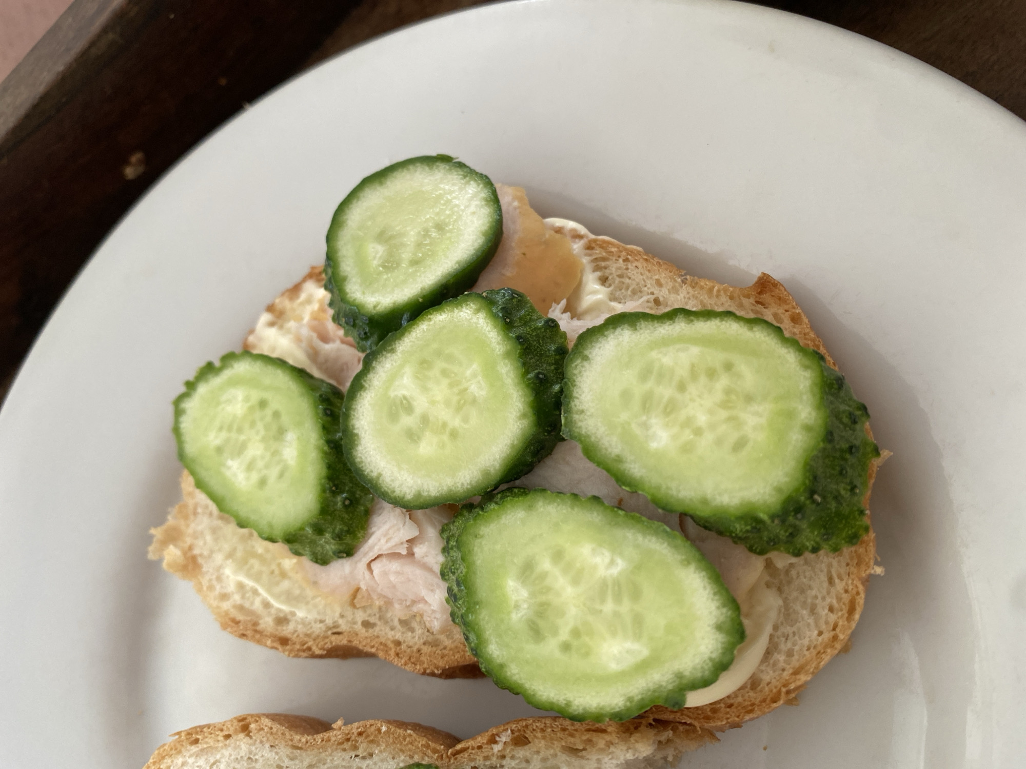 A close-up, overhead shot shows a sandwich on a white plate. The sandwich is made with a slice of bread, topped with a spread, possibly mayonnaise, and thin slices of what appears to be cooked meat. Five circular slices of cucumber are arranged on top of the meat. The sandwich is part of a larger, unidentifiable object on the left side of the frame, also appearing to be bread. The background is blurred, with a dark, wooden texture visible on the right. The scene is lit with natural light, creating a bright and clear image. This image was captured in the city of Chornomorsk, Ukraine.