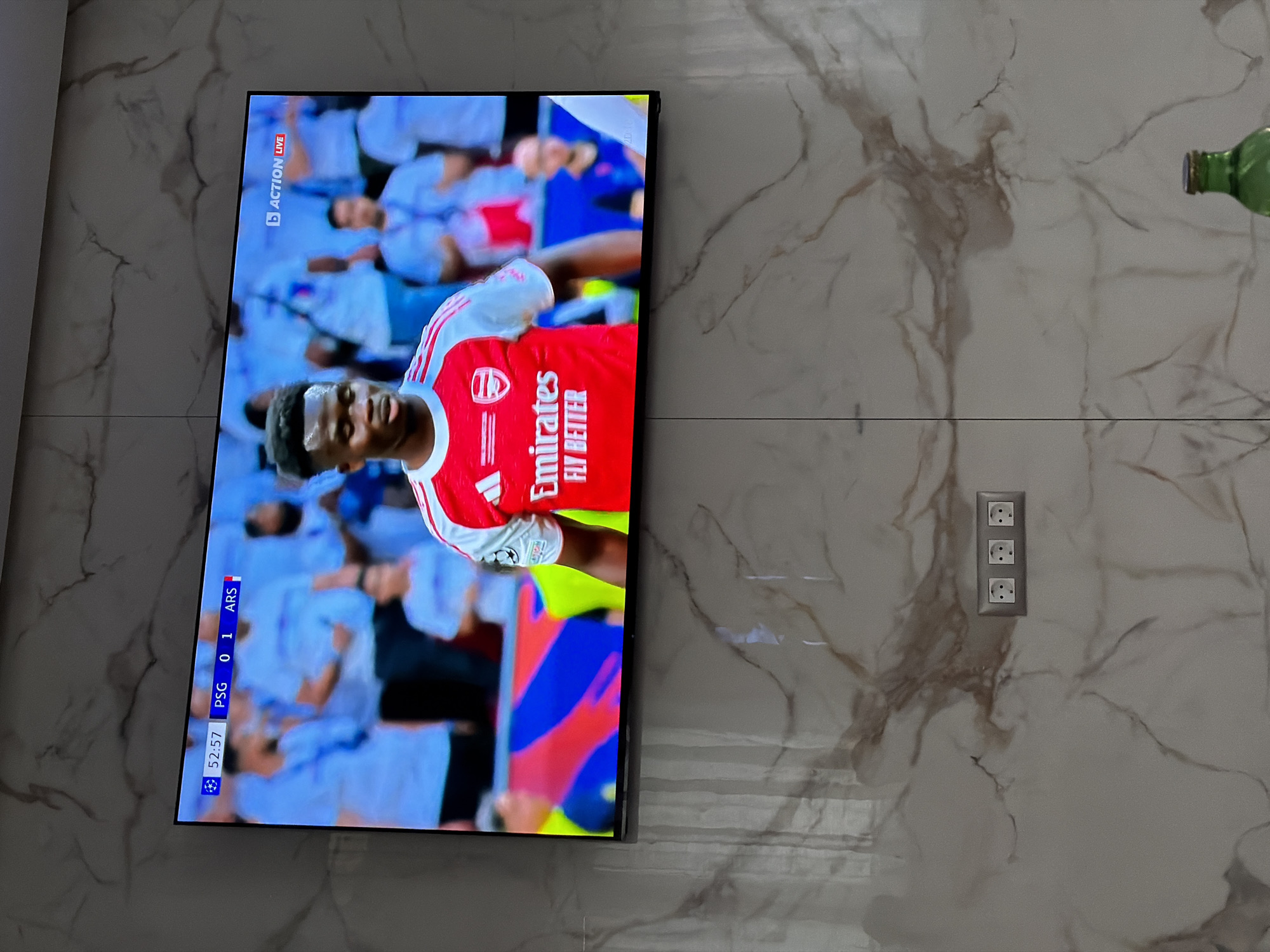 The image shows a flat screen television mounted on a marble wall in an interior space. The television screen displays a football (soccer) match, with a player in a red and white Arsenal jersey prominently featured. The score displayed on the screen shows PSG 0 and ARS 1. The match is live, as indicated by the "ACTION LIVE" logo.
Three electrical outlets are visible on the wall to the right of the television. A green bottle is partially visible in the upper right corner of the frame.
The location is not discernible from the visual information provided to infer its location in the city of Asenovgrad, Bulgaria.