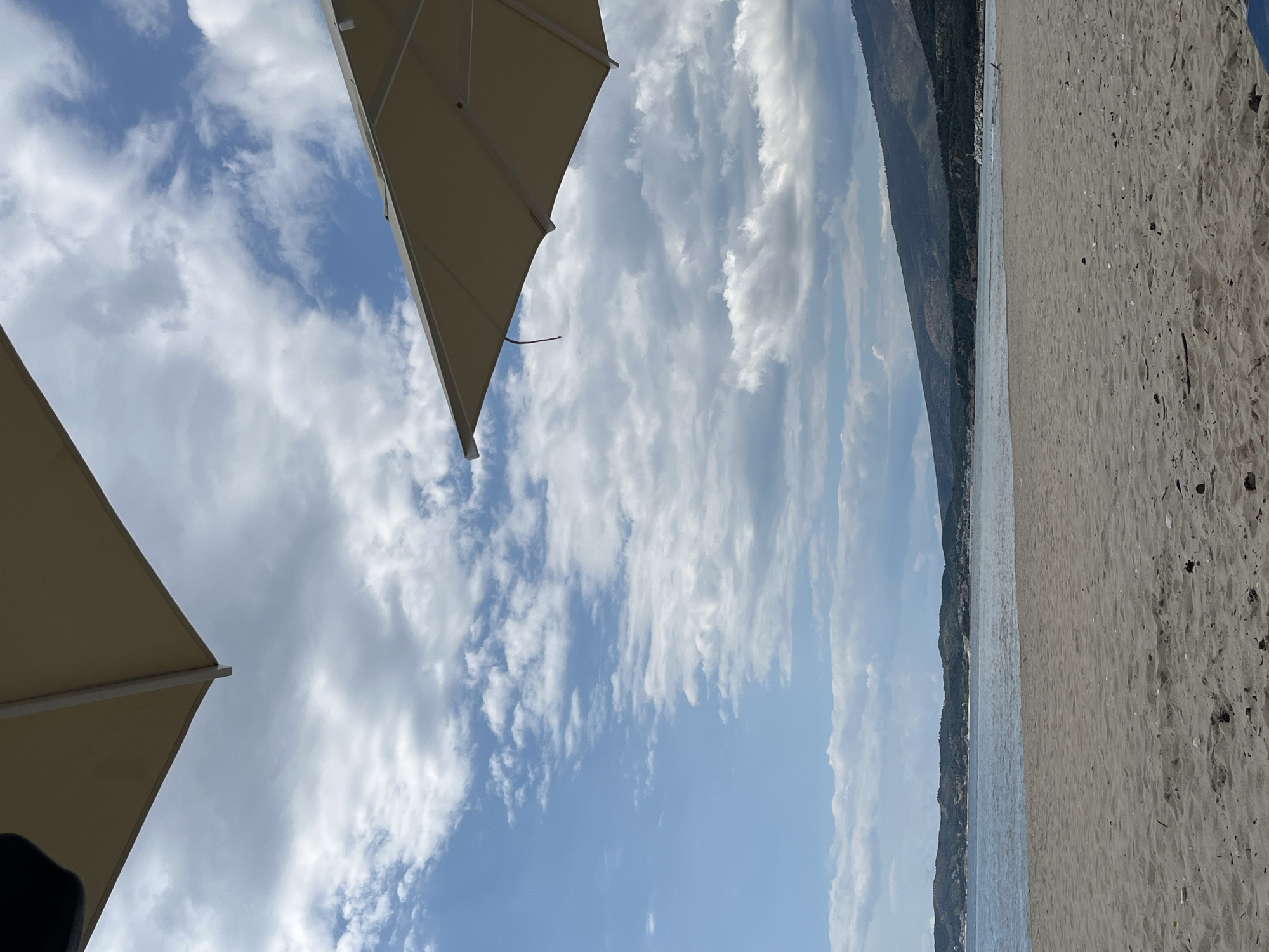 This is an eye-level, full shot of a beach with two umbrellas in the foreground. The beach is sandy with some scattered debris. The sea is calm and reflects the blue sky, which is dotted with white clouds. In the background, a mountain range covered in green vegetation slopes down to the sea. The scene is likely a beach resort or a public beach. The umbrellas suggest a place for relaxation and shade, implying a tourist or leisure destination.
