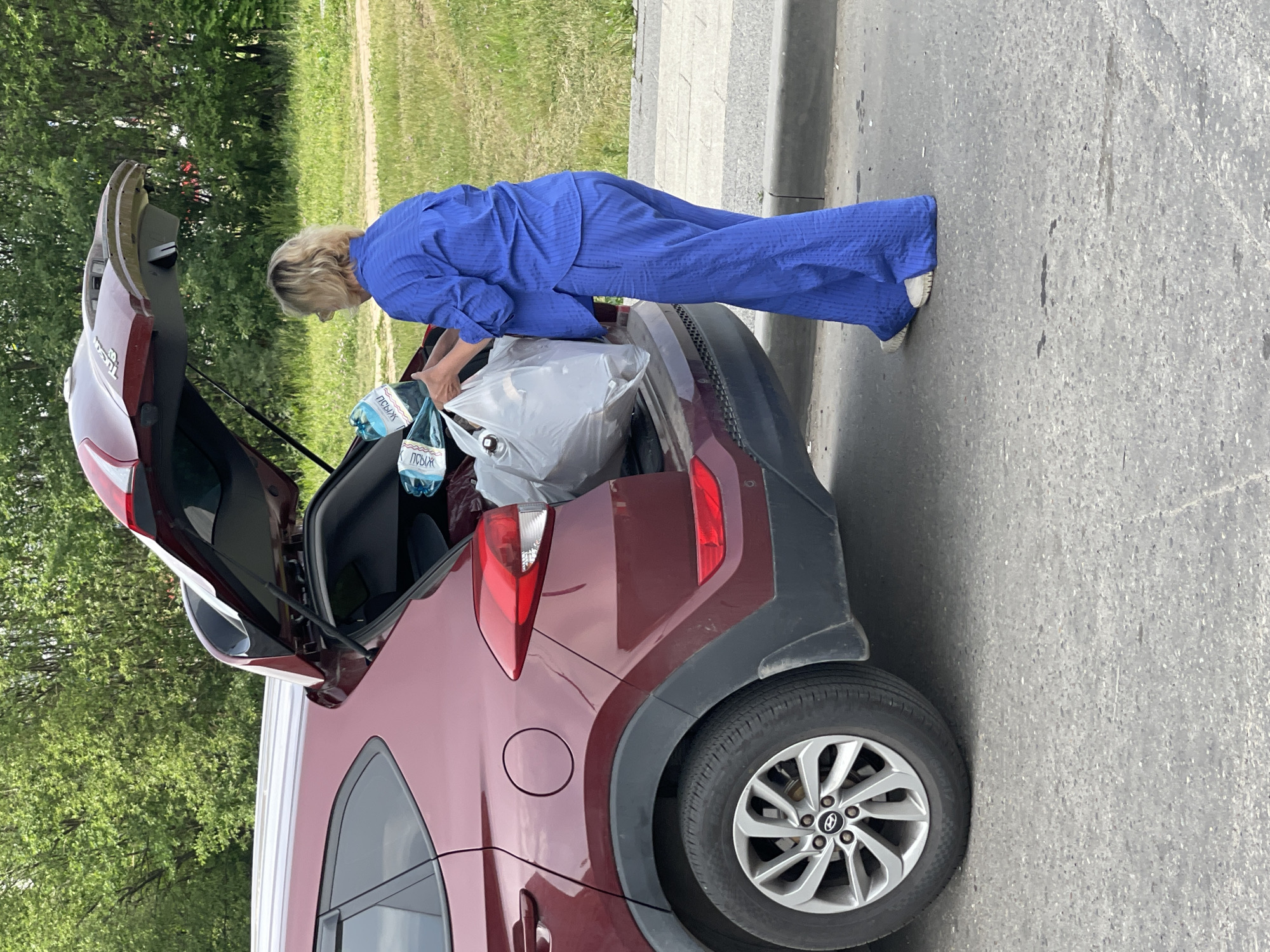 A blonde woman wearing a blue striped suit is standing next to the open trunk of a maroon Hyundai Tucson. She is holding a large white plastic bag containing bottles of water. The car is parked on a paved surface next to a grassy area with trees in the background. The scene appears to be a street or parking lot in Moskva, Russia.