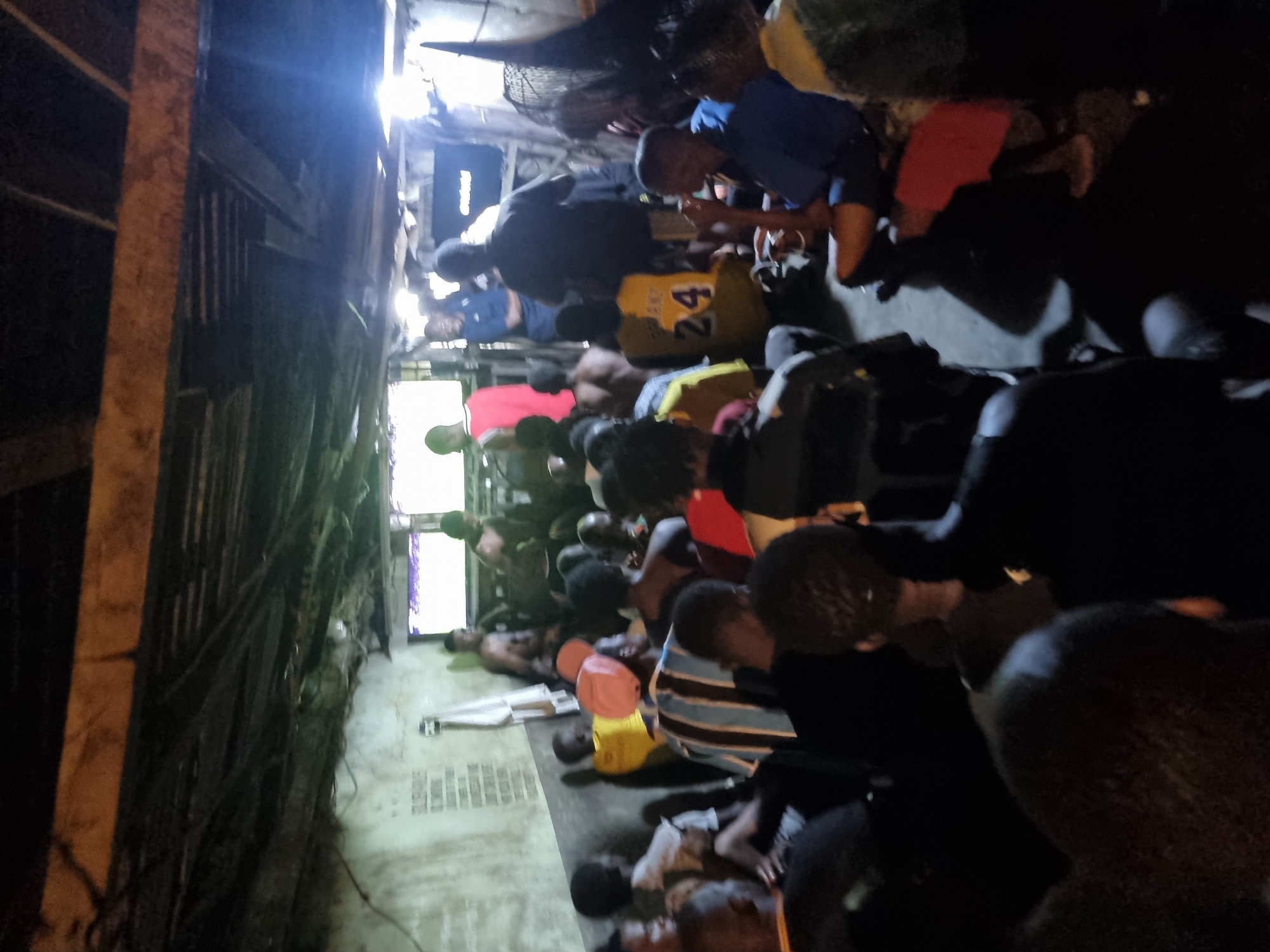 The image depicts a crowded room in what appears to be Onitsha, Nigeria. Numerous individuals, mostly young men, are seen lying or kneeling on the floor. Some are shirtless, while others wear various clothing items. There is an overhead fan and light fixtures visible, creating a dimly lit environment. The room's walls look bare and weathered. The scene suggests a confined space with limited resources.