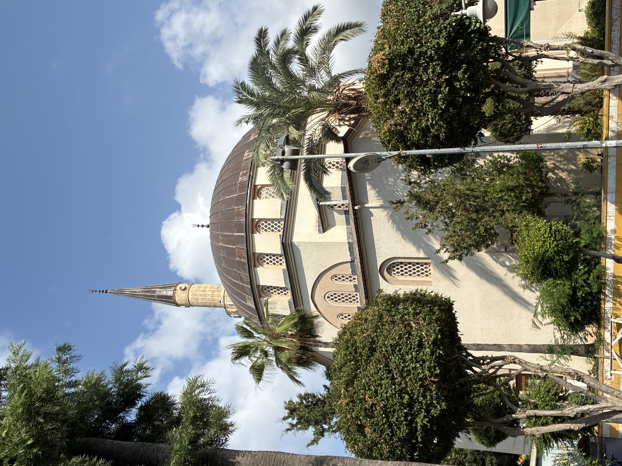 The image shows a mosque with a dome and a minaret, surrounded by trees and palm trees, under a bright blue sky with scattered clouds. The mosque has a beige exterior and features arched windows with decorative latticework. A street lamp is visible in the foreground, positioned in front of a palm tree. The overall scene is illuminated by natural daylight, casting shadows on the building and surrounding vegetation. This mosque is identified as the Emir Bedreddin Mosque, located in Alanya, Turkey. There are no people or animals visible in the image, and no actions or interactions are depicted.