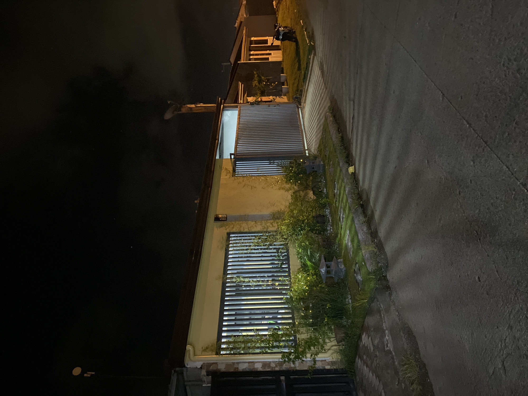 The image captures a residential street scene at night in Davao City, Philippines. A single-story house with a yellow facade and a dark-colored roof is prominently featured on the left. The house has a window covered by vertical metal bars, with greenery and decorative garden elements in front. A tall metal gate is also visible.
Adjacent to this house, another house with a white facade and a dark roof is seen. A motorbike is parked on the lawn in front of this second house. Further down the street, another house is visible with a similar architectural style.
The street itself is paved with asphalt and shows some wear and tear, with cracks and uneven patches. The scene is illuminated by artificial lights, casting long shadows across the road. The sky above is dark and appears overcast or cloudy. There are no people visible in the image. The overall impression is of a quiet residential neighborhood at rest during the night.