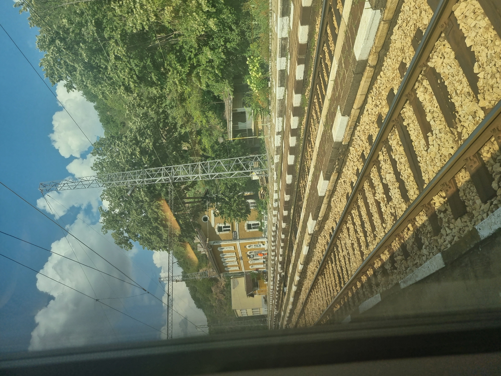 The image shows a view from a train window, looking out at a railway station in Zlidol, Bulgaria. The foreground is dominated by multiple sets of train tracks, with ballast visible between the wooden sleepers. Beyond the tracks, a low, yellow building with a red-tiled roof and white trim serves as the station building. It features several windows and a sign reading "ZLATOL". A single person in an orange uniform stands near the entrance of the building. Adjacent to the station is a smaller, older-looking wooden structure. Behind the buildings, a dense green forest covers a hillside. Overhead, numerous power lines crisscross the frame, supported by tall metal pylons. The sky is blue with scattered white clouds. The overall scene depicts a functional, somewhat rustic railway station set against a natural backdrop.