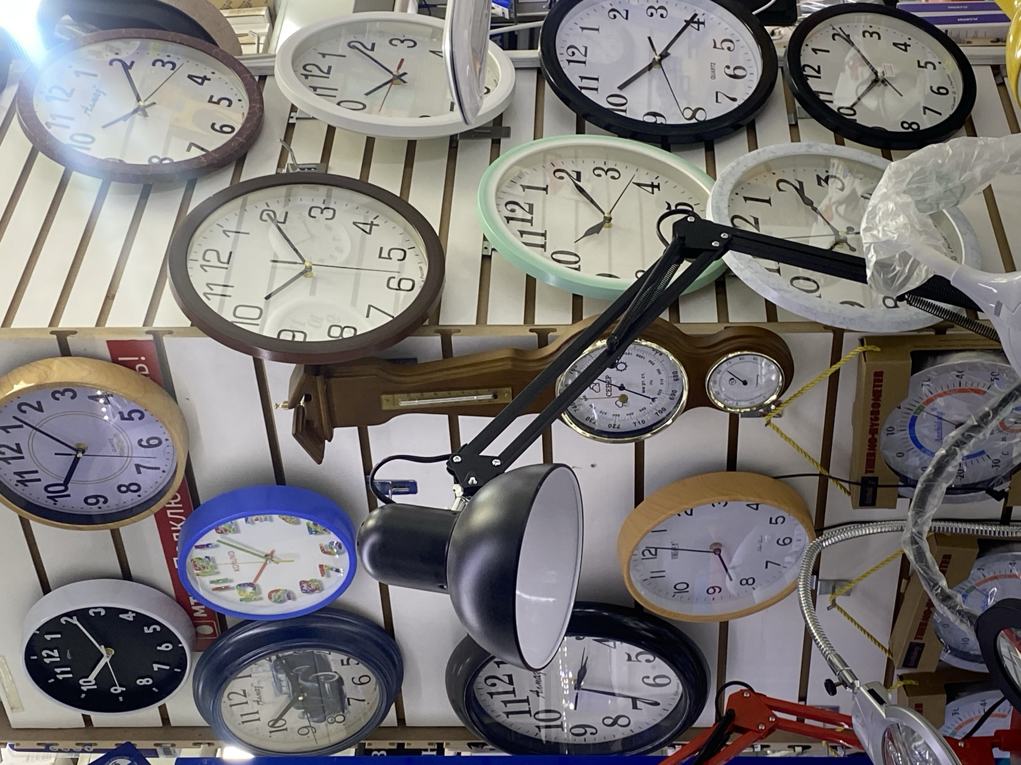 This media file captures a busy retail display, likely in a store in Moscow, Russia, showcasing a variety of wall clocks and other household items mounted on white slatwall panels. The scene is dominated by numerous round and oval clocks of various designs, colors, and sizes, suggesting a wide selection for customers. Many clocks display times around 10:10 or 10:45, a common aesthetic for merchandising.
Notable objects include a prominent black articulated desk lamp with a bell-shaped shade, positioned centrally, and a silver-colored flexible magnifying lamp with an illuminated head on the right side. A wooden weather station, featuring a barometer, thermometer, and hygrometer, is visible above the black lamp. Many clocks feature standard Arabic numerals, while one playful children's clock has colorful, distinct numbers. Some items in the background are partially obscured or wrapped in clear plastic. A bright light source in the upper left corner causes significant glare on some of the clock faces.
Visible text on the clocks and instruments includes brand names such as "QUARTZ", "Acuag", "KORUND", "Anzhao", "INTEST", "Humay", and "CEBEP". The weather station has "HYGROMETER" and "мм рт. ст." (millimeters of mercury) inscribed, indicating its functions. Other partially visible text includes "HUAYU" and "MUAYM" on packaging or labels. The Cyrillic text further supports the given location context of Moscow, Russia. The image provides a snapshot of a diverse inventory, likely from an electronics or home goods store.