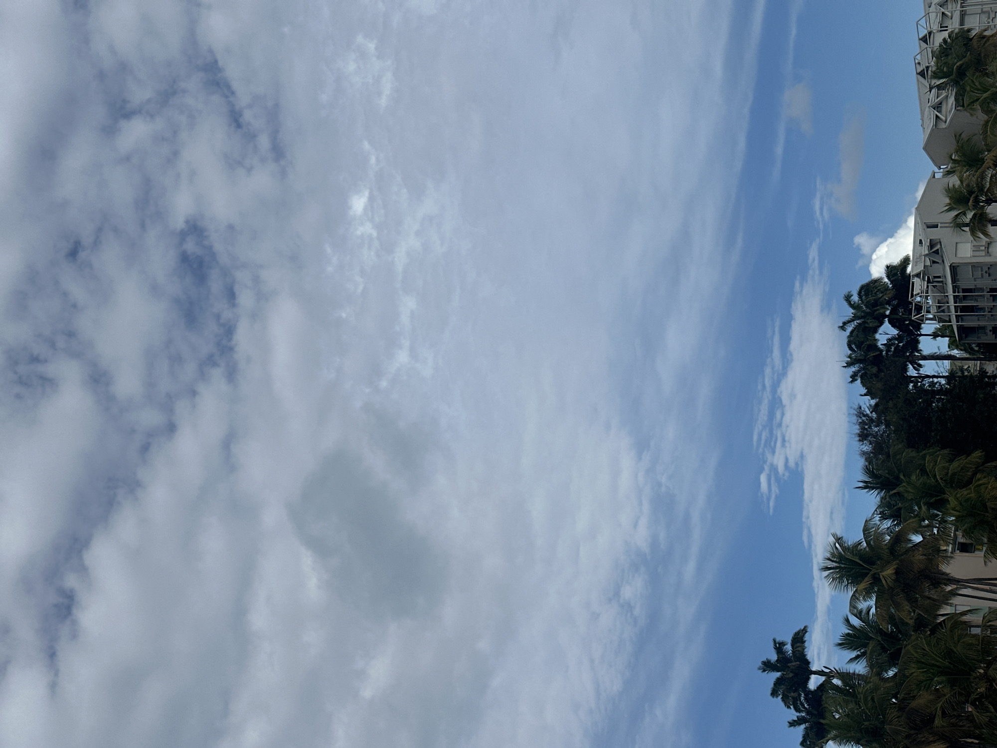 The image depicts a sky with scattered clouds over Le Gosier, France. Palm trees are visible on the right side of the frame, partially obscuring a multi-story building with white balconies. The sky is predominantly blue, with patches of dense, white clouds.