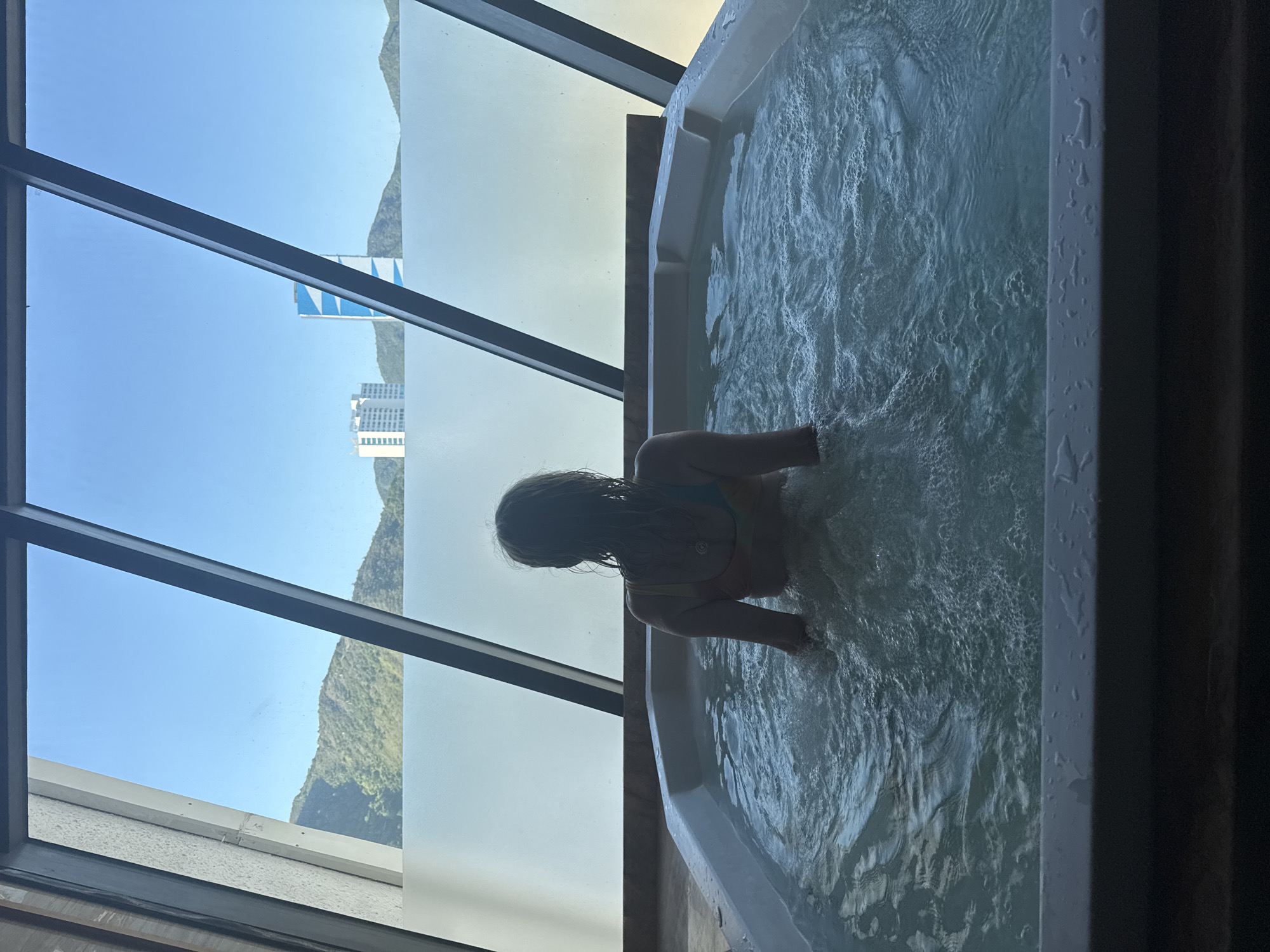 In a hotel room in Nha Trang, Vietnam, a woman enjoys a hot tub. The room features a large window with a sloped glass ceiling. Through the window, the city of Nha Trang is visible, with distant green mountains and modern buildings. The woman, seen from behind, is wearing a yellow and blue bikini. She is immersed in the bubbling water of the hot tub, which is partially obscured by steam and the surrounding glass. The scene suggests a relaxing and luxurious experience, with the city view providing a scenic backdrop.