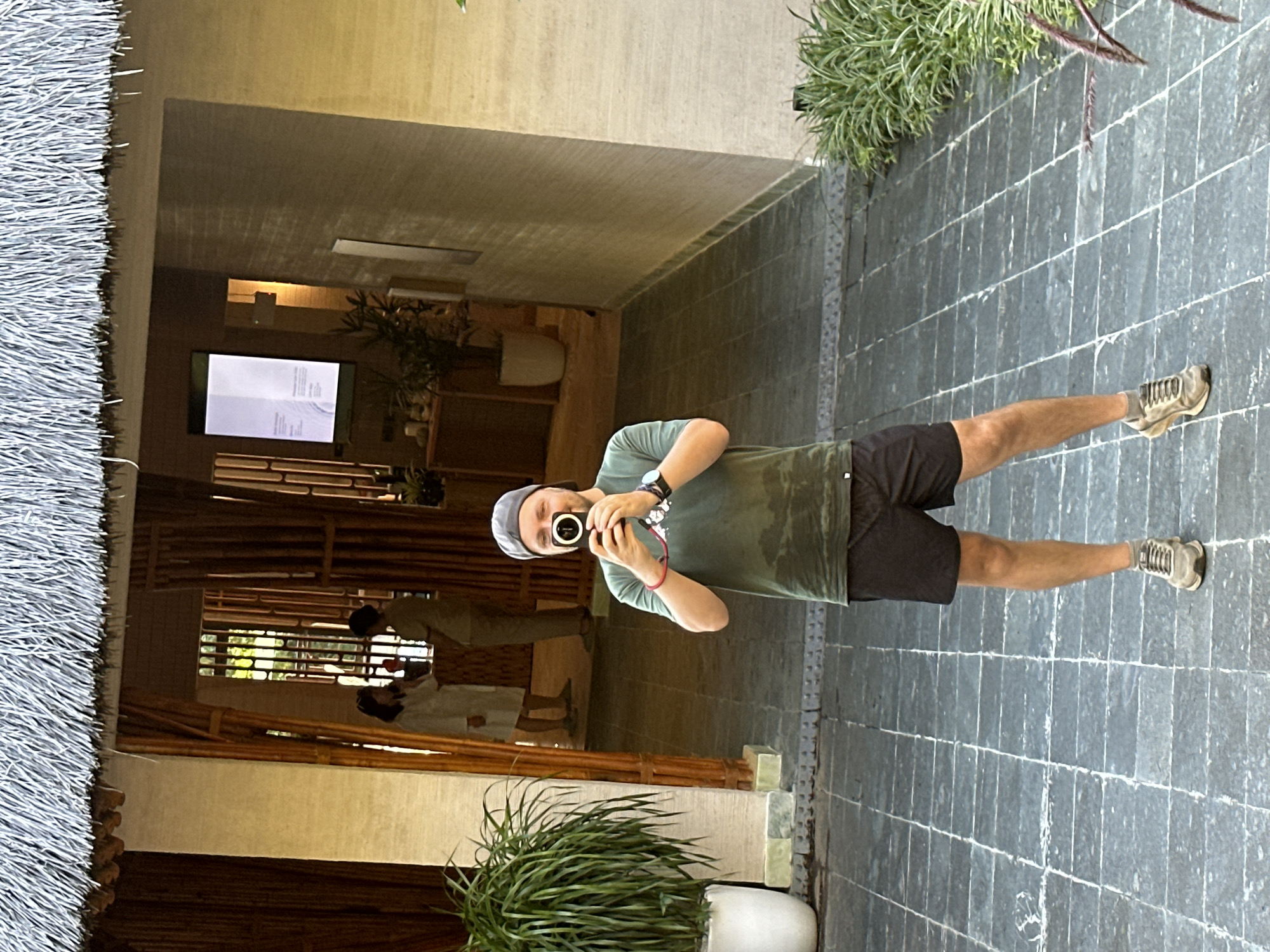 The image shows a man taking a mirror selfie in what appears to be a resort or hotel setting in Mu Cang Chai, Vietnam. The photograph is oriented vertically, with the man's body appearing horizontal across the frame.
The central subject is a man, wearing a green short-sleeved t-shirt, black shorts, a grey baseball cap, and light-colored sneakers. He is holding a black digital camera, pointing it towards the reflective surface to capture his own image. A watch is visible on his left wrist, along with a red bracelet.
The background reveals an inviting indoor-outdoor space. To the left, a textured thatched roof creates a natural, rustic architectural element. A dark grey tiled pathway or floor with distinct white grout lines stretches across the lower portion of the image. Potted green plants are strategically placed, enhancing the natural ambiance; one large plant in a white pot is prominent in the foreground.
Beyond the man's reflection, the interior space features warm-toned walls and intricate bamboo structures, suggesting traditional Vietnamese design. In the deeper background, two blurry figures are subtly visible, possibly reflections of other people or actual individuals within the space. A rectangular screen or monitor mounted on a wall displays light-colored text, which is illegible due to distance and resolution, but suggests an information display or menu.
The lighting indicates it is daytime, casting a bright but diffused light over the scene. The overall atmosphere is calm and peaceful, characteristic of a serene holiday destination.