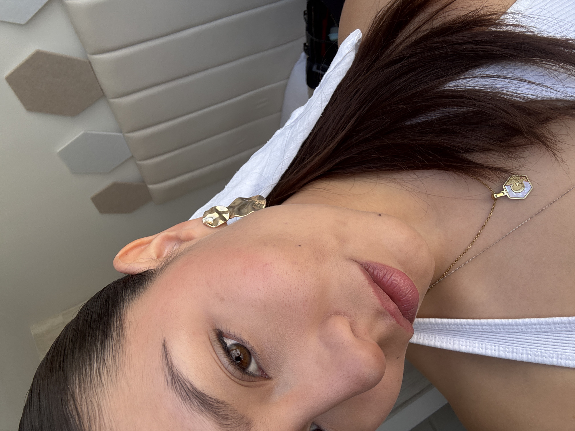 A close-up shot of a woman's face, turned at an angle. She has dark hair, brown eyes, and is wearing a white top. She also has a gold earring and a gold necklace. The background includes a textured wall. The location is not discernible from this image.