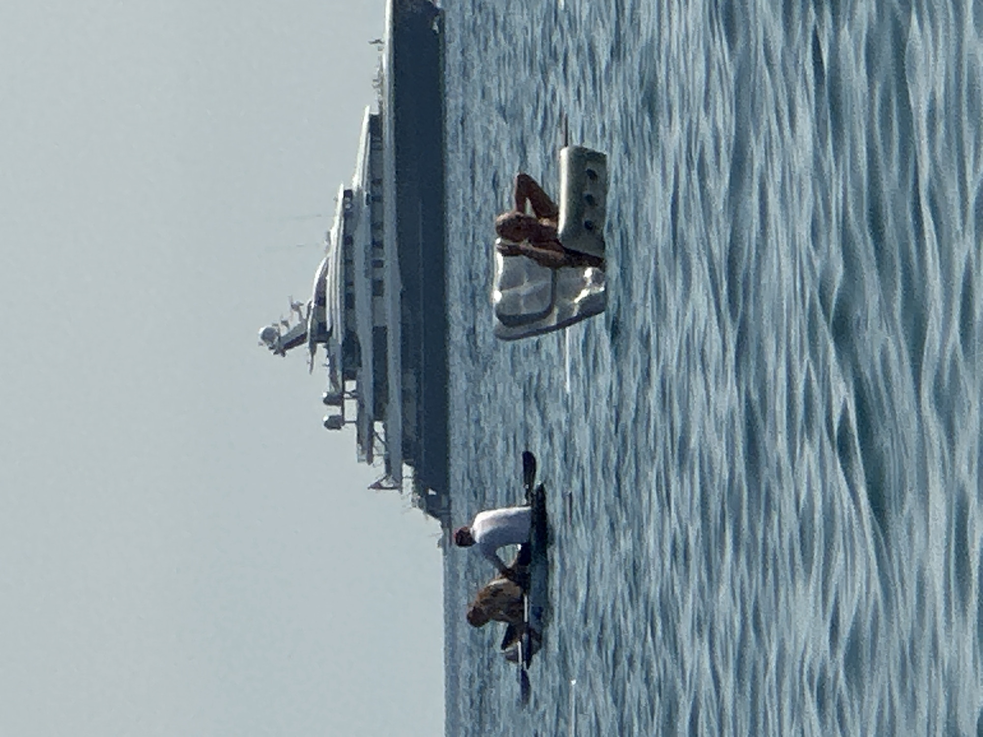 In the waters off the city of Boreti, Montenegro, a large, dark-hulled yacht is anchored. In the foreground, two people are in a small boat. One person is seated at the front of the boat, facing away from the camera. The other person is seated behind them, facing towards the camera and appears to be paddling. To the right of this boat, a person is reclining in a silver, boat-like object, with their hands behind their head. The water is choppy, with visible ripples. The sky is a pale, hazy blue.