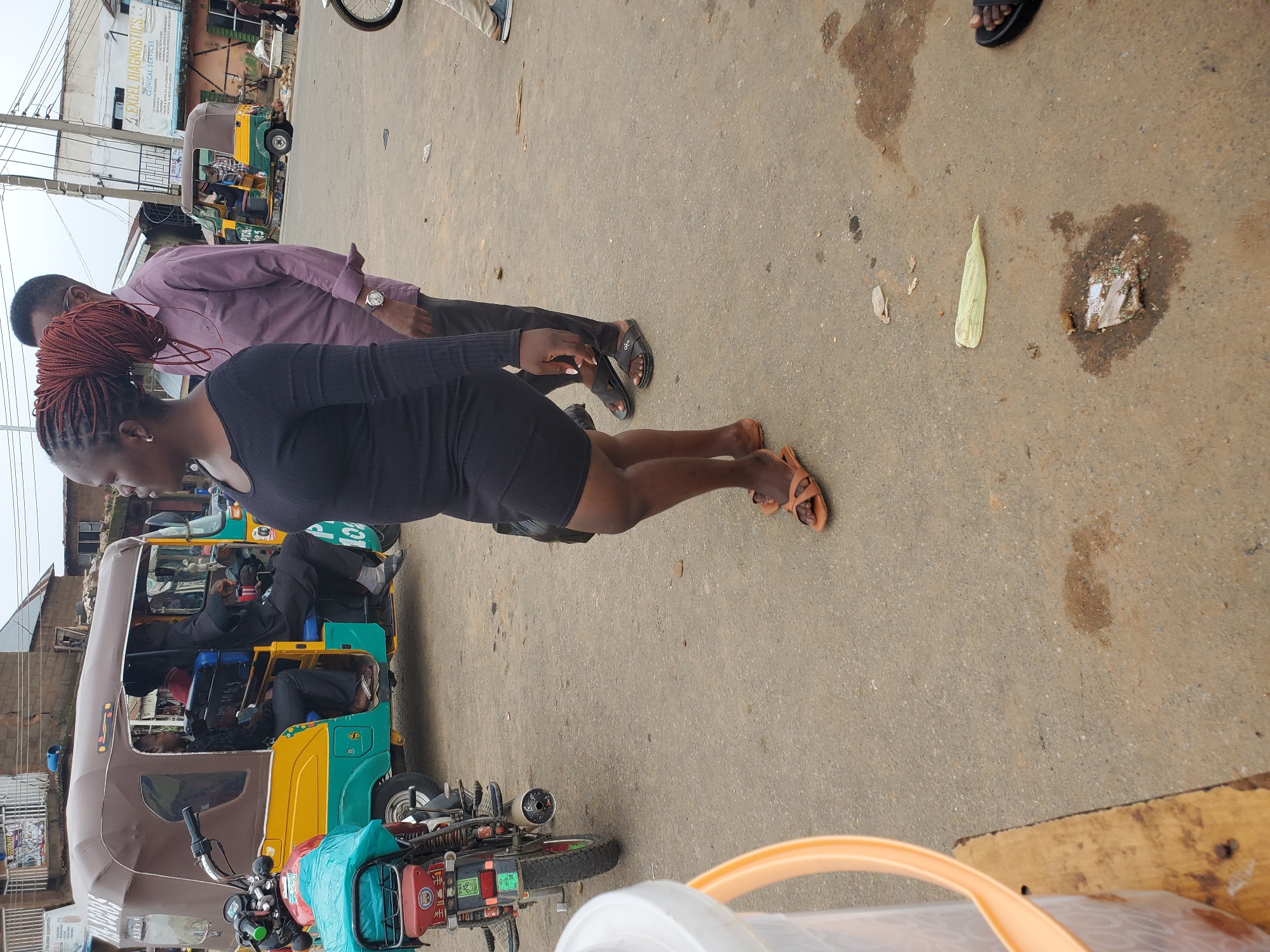In Gwong, Nigeria, on a paved street, a Black woman with braided hair stands on a sidewalk next to a tricycle. The woman is wearing a black dress and brown sandals. To her left, a man in a purple shirt and black pants stands. Behind them, the tricycle is yellow, green, and brown, and it is carrying several passengers. The street is lined with buildings and shops. The environment appears to be a typical urban street scene.