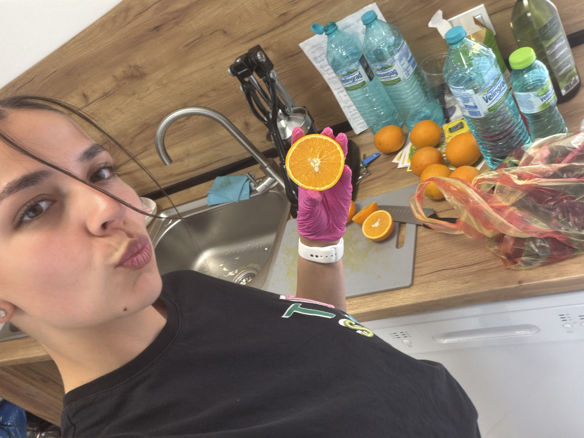 A young woman is taking a selfie in what appears to be a kitchen. She is holding half of an orange in a pink gloved hand. The kitchen counter features a sink, faucet, bottles of "Velingrad" mineral water, oranges, a cutting board, a knife, and a grocery bag. The woman has dark hair and is wearing a dark t-shirt. A white dishwasher is located below the counter. While the image doesn't definitively establish the location as Gabrovo, Bulgaria, the presence of "Velingrad" mineral water, a brand common in Bulgaria, suggests a possible connection to the region.