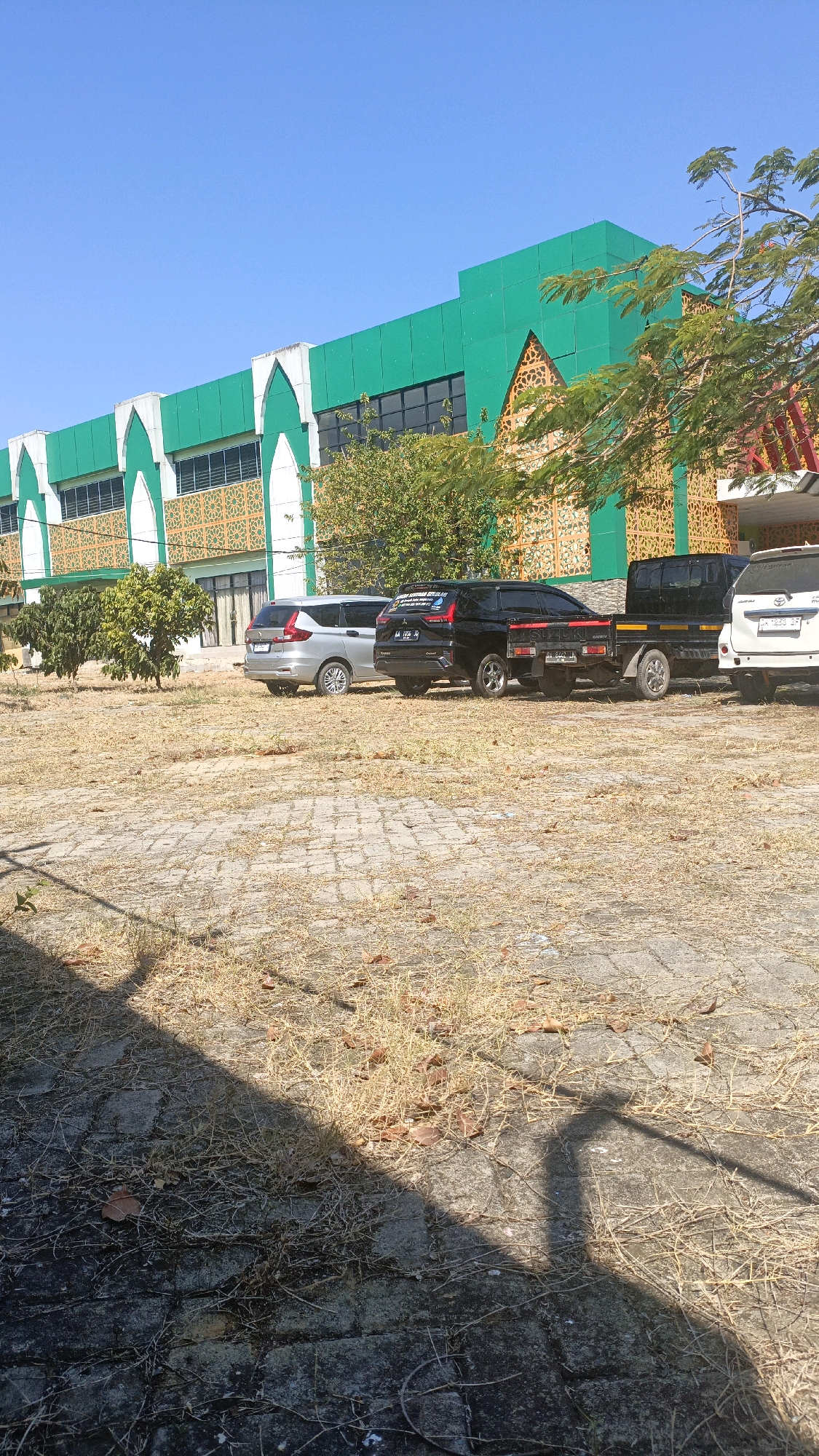 The image shows the exterior of a modern building with green and white arched architectural elements, reminiscent of Islamic design, in Desa Pabahanan, Pelaihari, Tanah Laut, Indonesia. The building features ornate golden latticework between the arches. In front of the building, there is a dry, grassy area with a paved ground made of interlocking stones. Several vehicles are parked on the paved area, including a silver MPV, a black SUV, a black pickup truck with a flatbed, and a white SUV. The sky is clear and blue, indicating a sunny day. The overall scene depicts a public or institutional building in an outdoor setting.