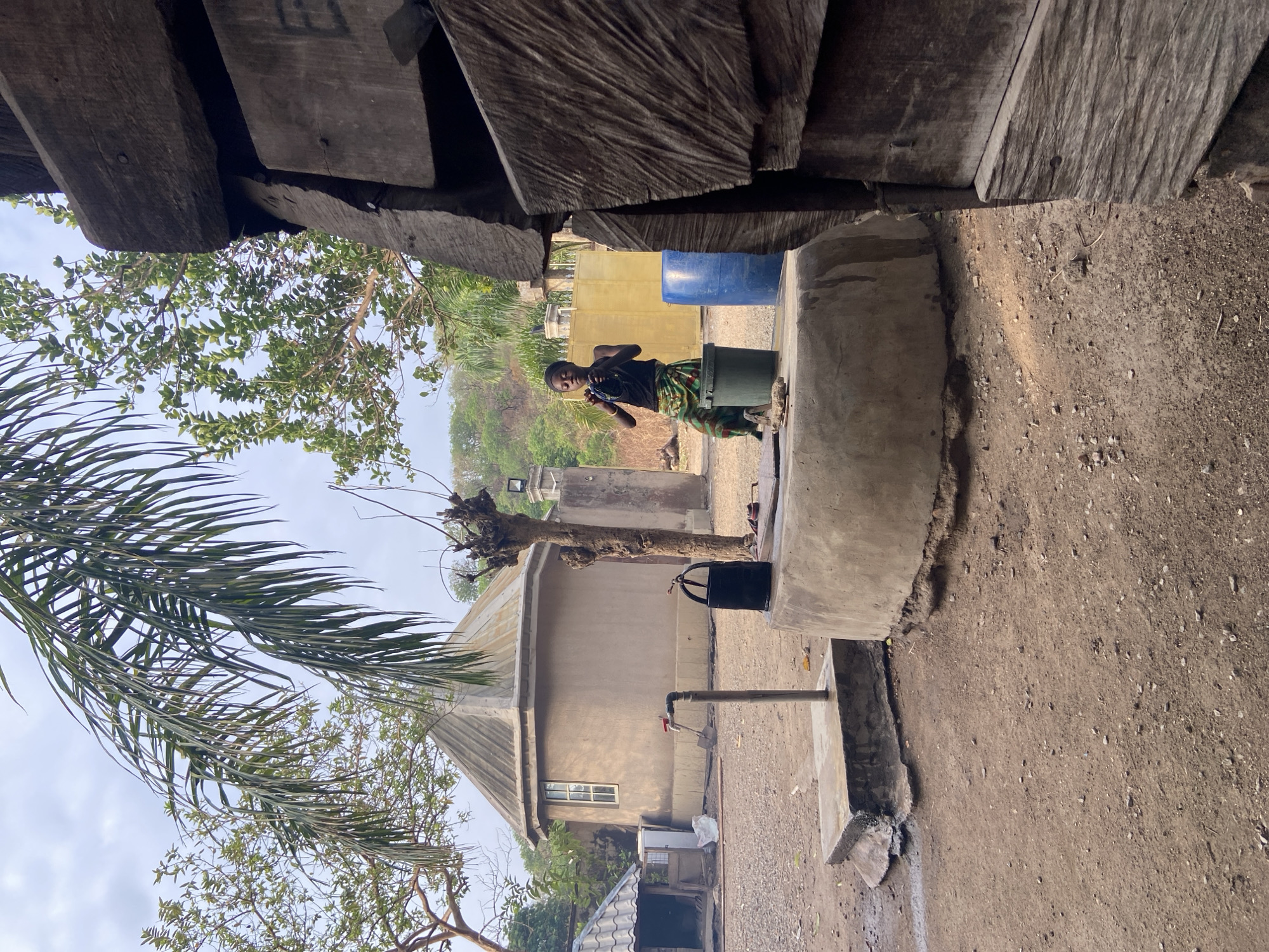 An exterior shot reveals a dirt ground surface and a young person of indeterminate gender standing atop a green container. The individual wears a black shirt and a wrap skirt with green and brown tones. They are holding an object and appear to be engaged with it. A blue plastic barrel is placed behind them. The setting appears to be an outdoor compound, possibly in Mararaba, Nigeria, with simple buildings and trees in the background. A dark bucket hangs from a pipe near the house. The photo is taken from underneath wooden beams which partly obstruct the view.