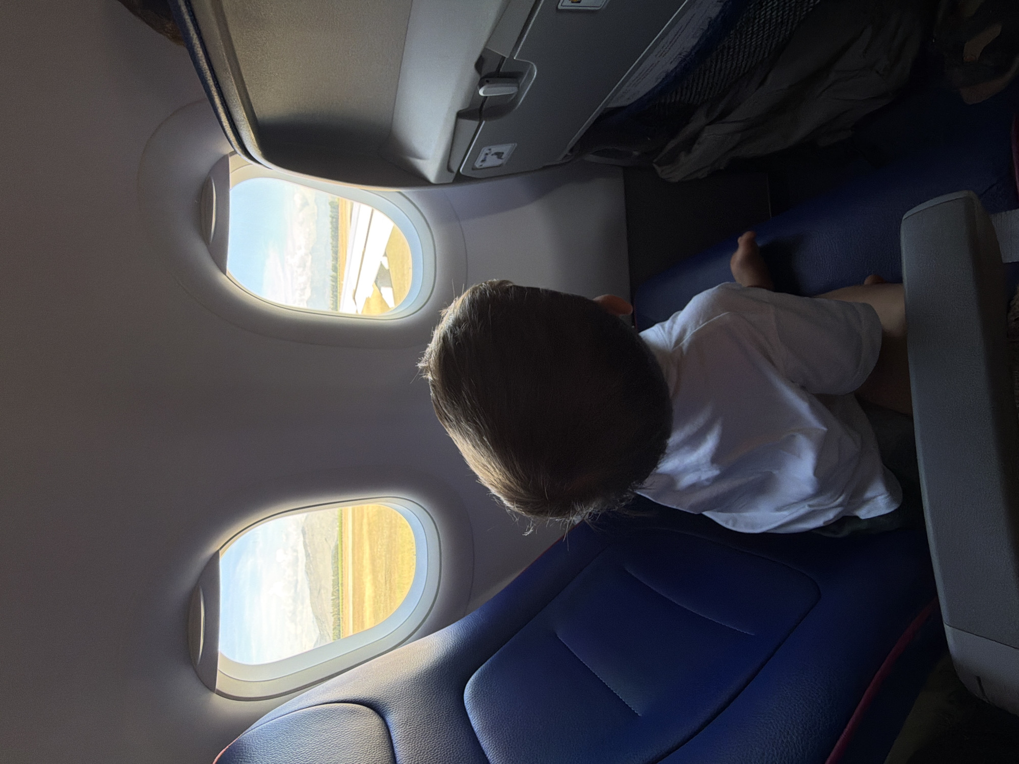 An overhead interior shot shows a child's head and shoulders as they look out of an airplane window. The child, wearing a white shirt, is seated in a blue airplane seat. The view outside the window reveals a sunny landscape with a mountain range in the distance, a tan-colored field, and the wing of the airplane. The airplane interior includes gray seatbacks with storage compartments visible. The image is not specific to any location in Donja Cijevna, Montenegro; it depicts a scene inside an airplane.