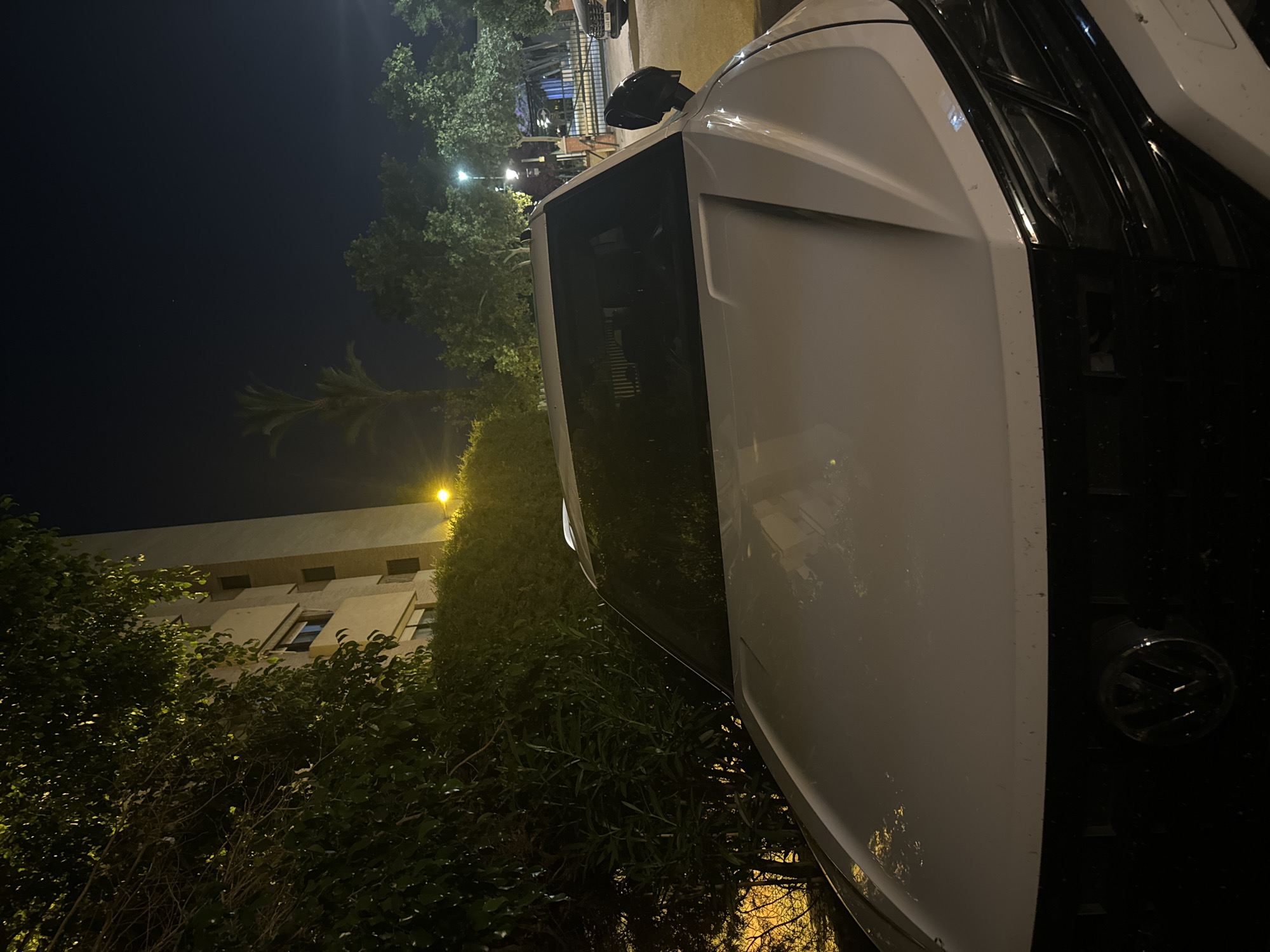 A white Volkswagen SUV is parked at night in front of a multi-story building, partially obscured by dense foliage. The scene appears to be in a residential area, as indicated by the building and street lighting. The car's headlights are not illuminated. The ground is paved and appears to be a street or parking area. There are trees and other greenery in the background, as well as a wrought-iron gate in the distance. The overall lighting suggests it is nighttime, with artificial light sources creating shadows and highlights.