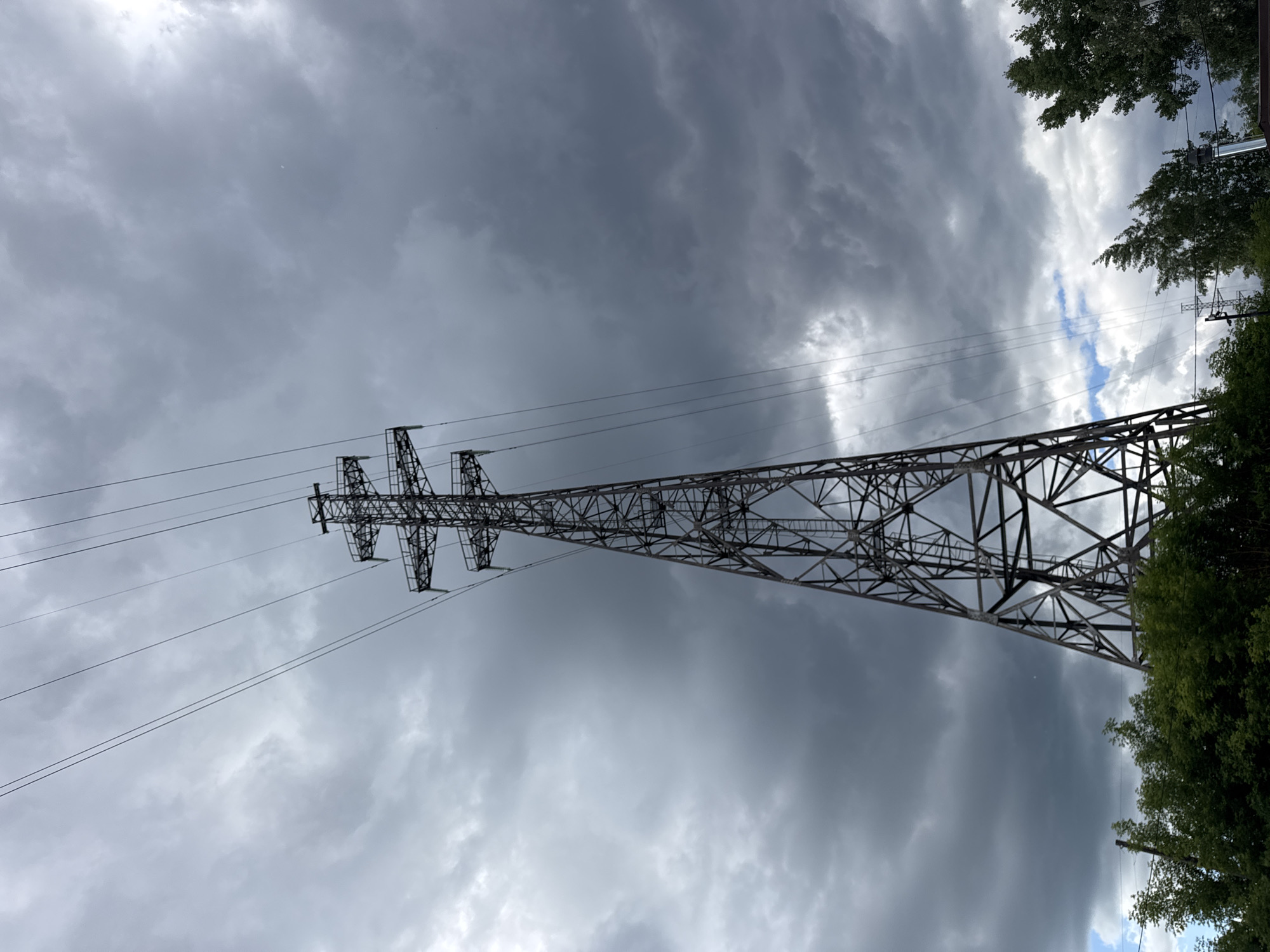 An electric pylon stands tall against a dramatic, cloudy sky. The pylon, made of dark metal lattice work, tapers upwards to support multiple high-voltage power lines that stretch across the frame. Below the pylon, a dense line of lush green trees forms the lower border of the image. The sky is filled with dark, heavy clouds, suggesting an impending storm or recent rain, with only hints of brighter sky visible through breaks in the cloud cover. The scene is devoid of people and any specific identifiable structures, giving it a somewhat stark and industrial feel, yet grounded by the presence of nature. The location is the city of Chaikovsky, Russia.