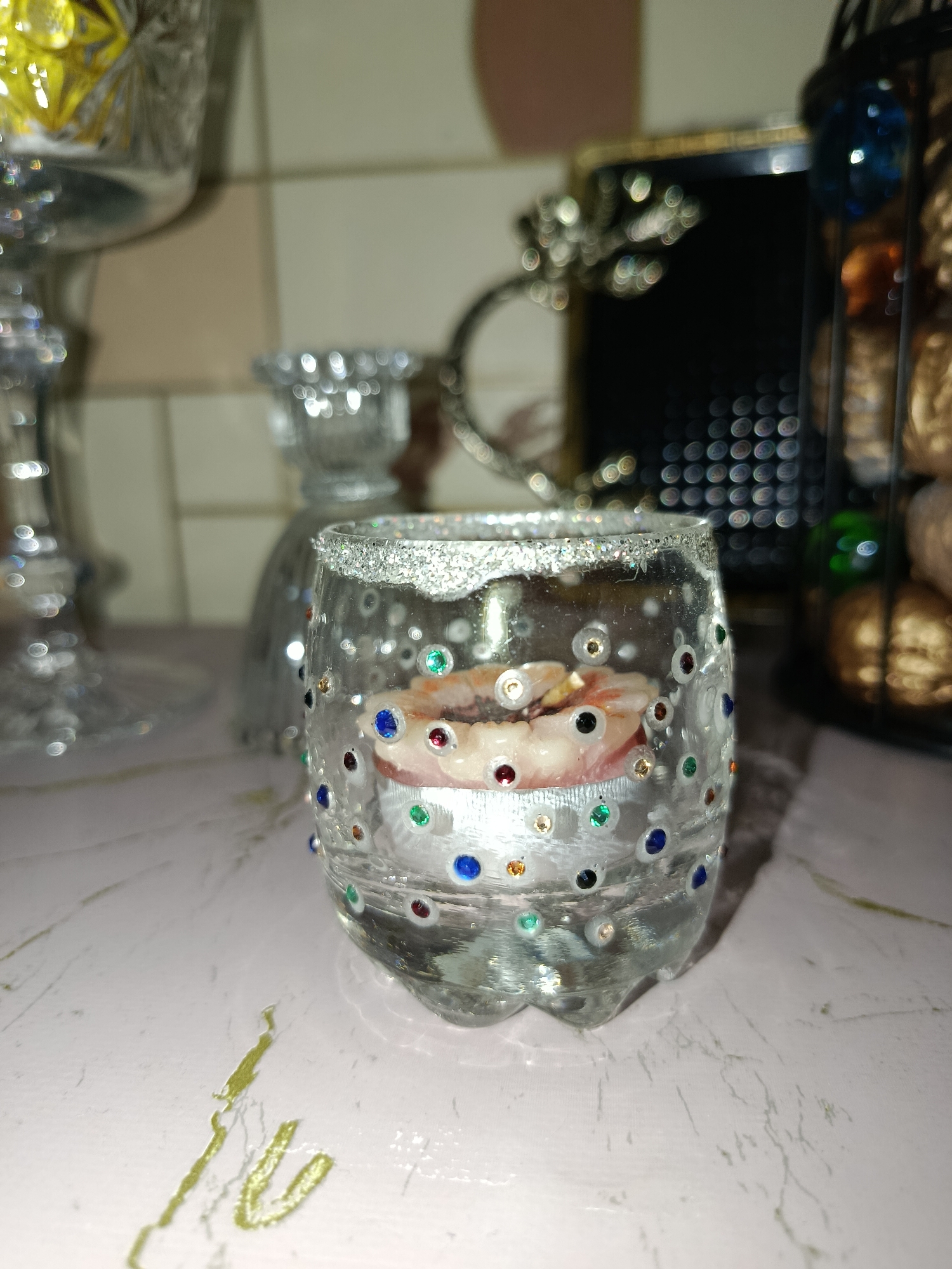In a domestic indoor setting, a close-up shot focuses on a clear glass votive holder, decorated with numerous colorful rhinestones and glitter around its rim. Inside the holder, a small, pink-colored candle, shaped like a flower, is visible. The background is blurred but suggests a kitchen or dining area with other decorative items, including a crystal goblet on the left and a wire basket containing what appear to be nuts on the right. The lighting is artificial, casting highlights and reflections on the surfaces. The scene is not identifiable as being in Tashkent, Uzbekistan.