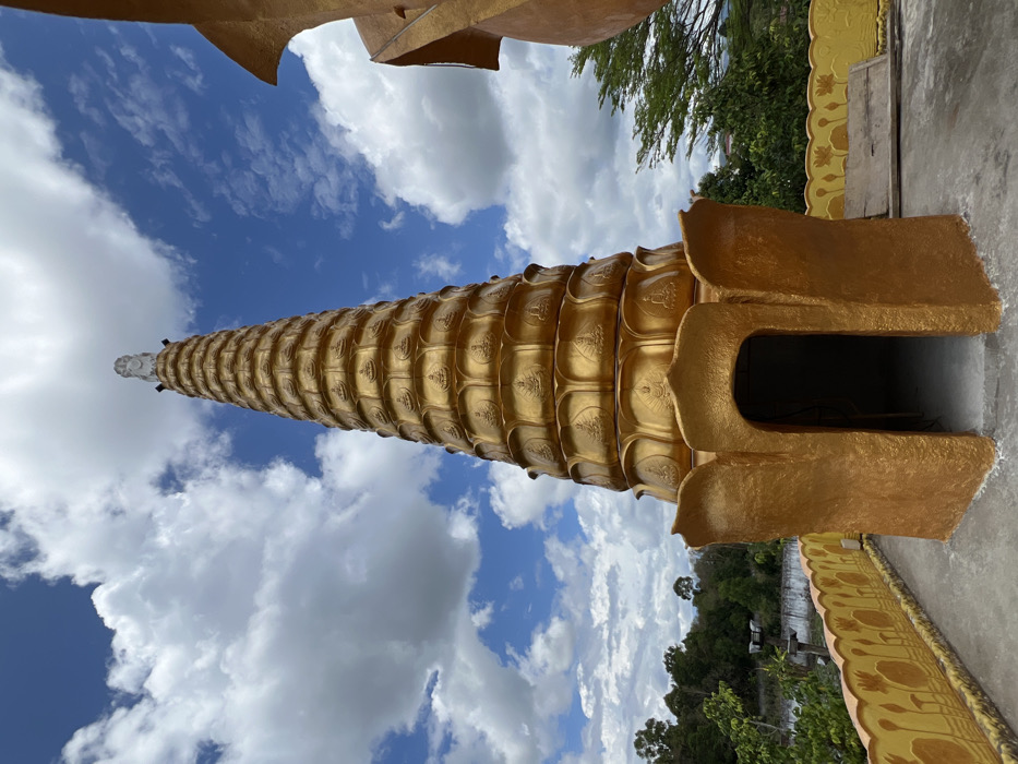 A golden, tiered stupa dominates the scene under a partly cloudy blue sky, located in Đăk Mar, Vietnam. The conical structure is adorned with multiple horizontal bands, each featuring a repetitive pattern of seated Buddha figures in relief. A white, sculpted figure crowns the apex of the stupa. At its base, a prominent golden structure with lotus-petal motifs forms an arched entrance into a darkened interior. The stupa stands on a flat, grey concrete platform. To the left, a low yellow wall or railing with stylized orange lotus designs extends along the platform's edge. On the far right, a curved golden architectural element of an adjacent building is partially visible. The background features lush green trees and distant vegetation, indicative of a natural, possibly elevated, setting. No individuals or specific actions are depicted.