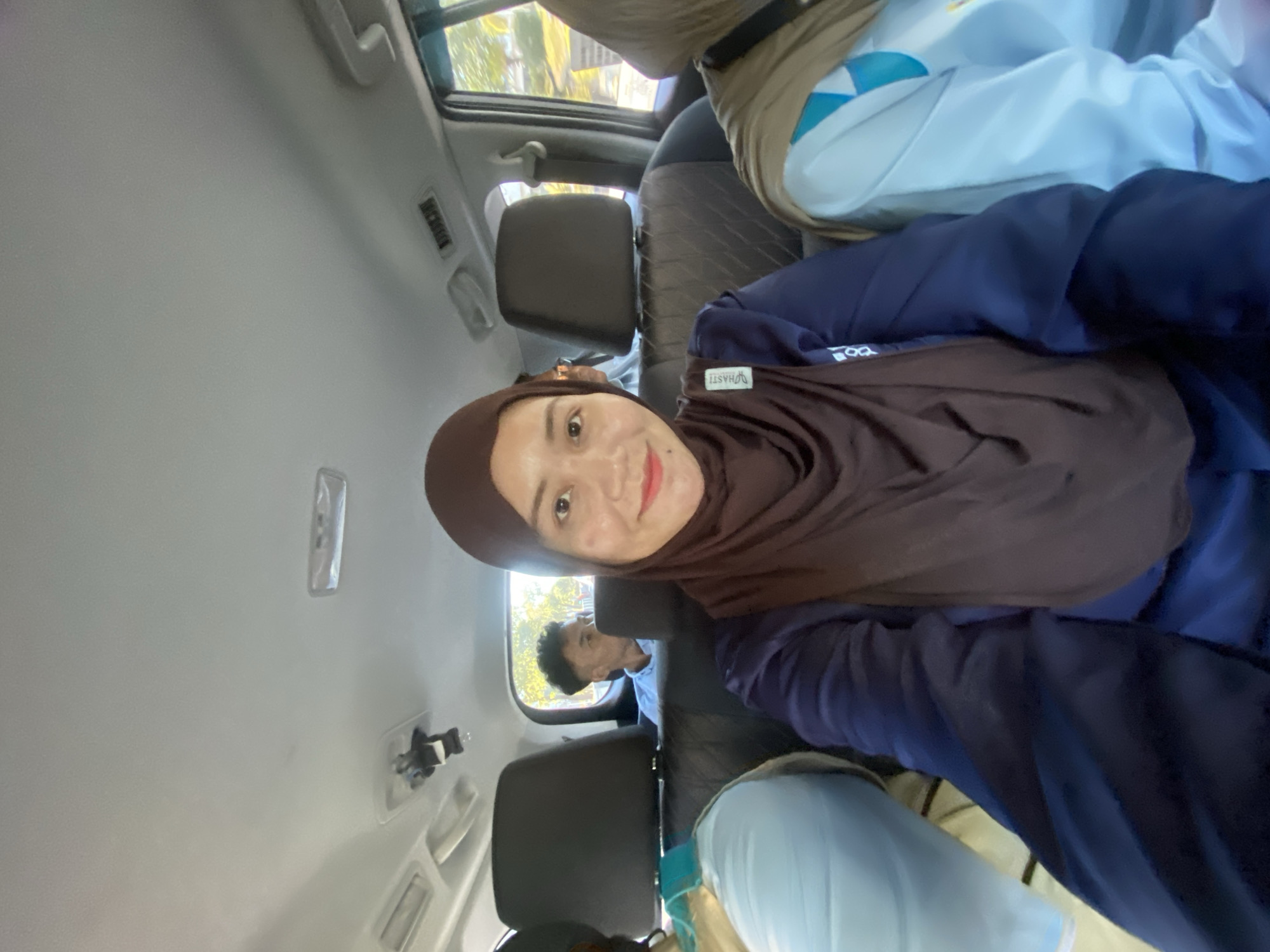 In a vehicle, likely a car given the interior configuration, a woman wearing a brown hijab and a navy blue long-sleeved shirt is taking a selfie. She is in the foreground, facing the camera with a slight smile. Behind her, a man in a light blue collared shirt is visible in the back seat, looking to the side. To the woman's left and right are other passengers, also wearing hijabs, with their heads and shoulders visible. The interior of the vehicle includes dark-colored headrests and ceiling lights. The scene suggests a journey, possibly within Banjarmasin, Indonesia, given the typical mode of transport and attire.