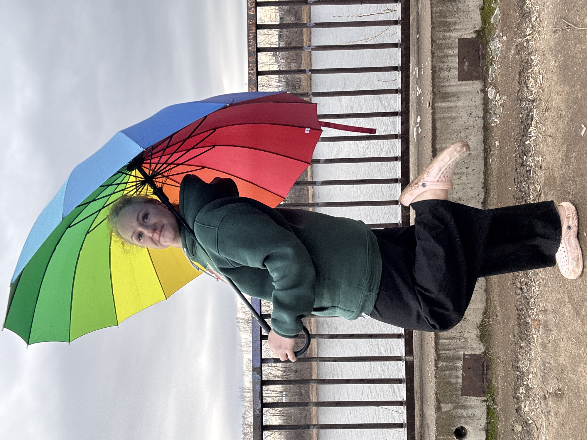 A young woman stands next to a metal railing on a concrete surface holding a rainbow-colored umbrella. She wears a green hooded sweatshirt and black pants. Her footwear consists of light pink clog-style shoes. The background reveals a body of water beyond the railing and a cloudy sky. The location appears to be in Chaikovsky, Russia, indicated by the inscription "Чайковский, Россия" visible on the railing. There is scattered debris on the ground near the railing.